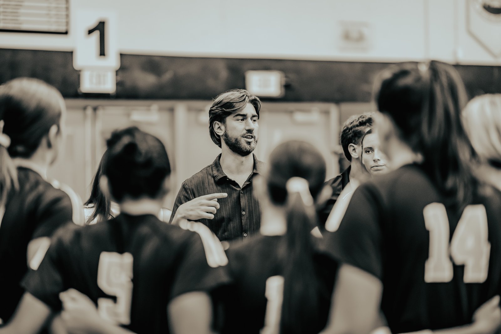 A coach gestures to players in black jerseys with white numbers, including 14, during a huddle on the court.