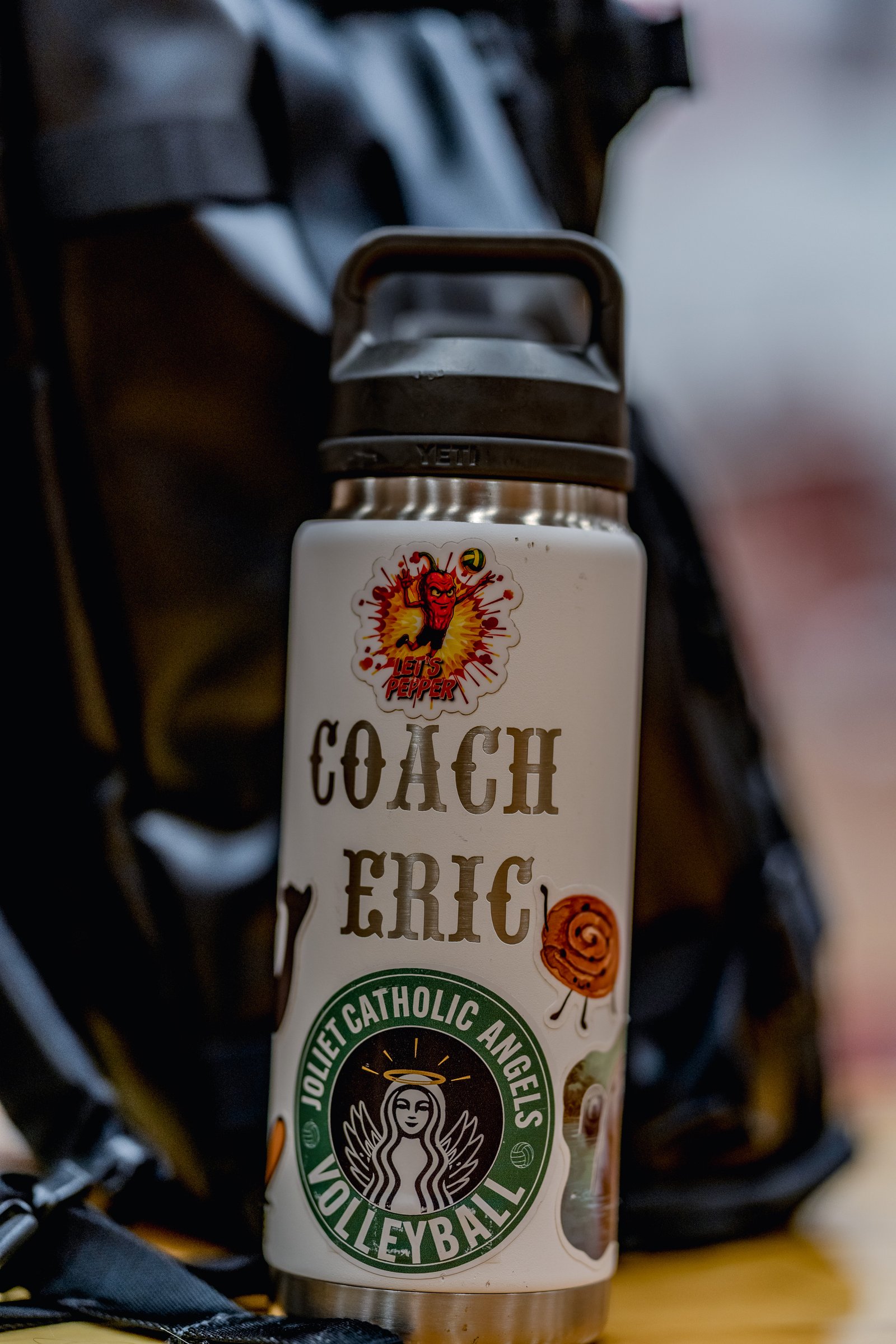 A water bottle with stickers for "Coach Eric" and "Joliet Catholic Angels Volleyball" sits near a black backpack.