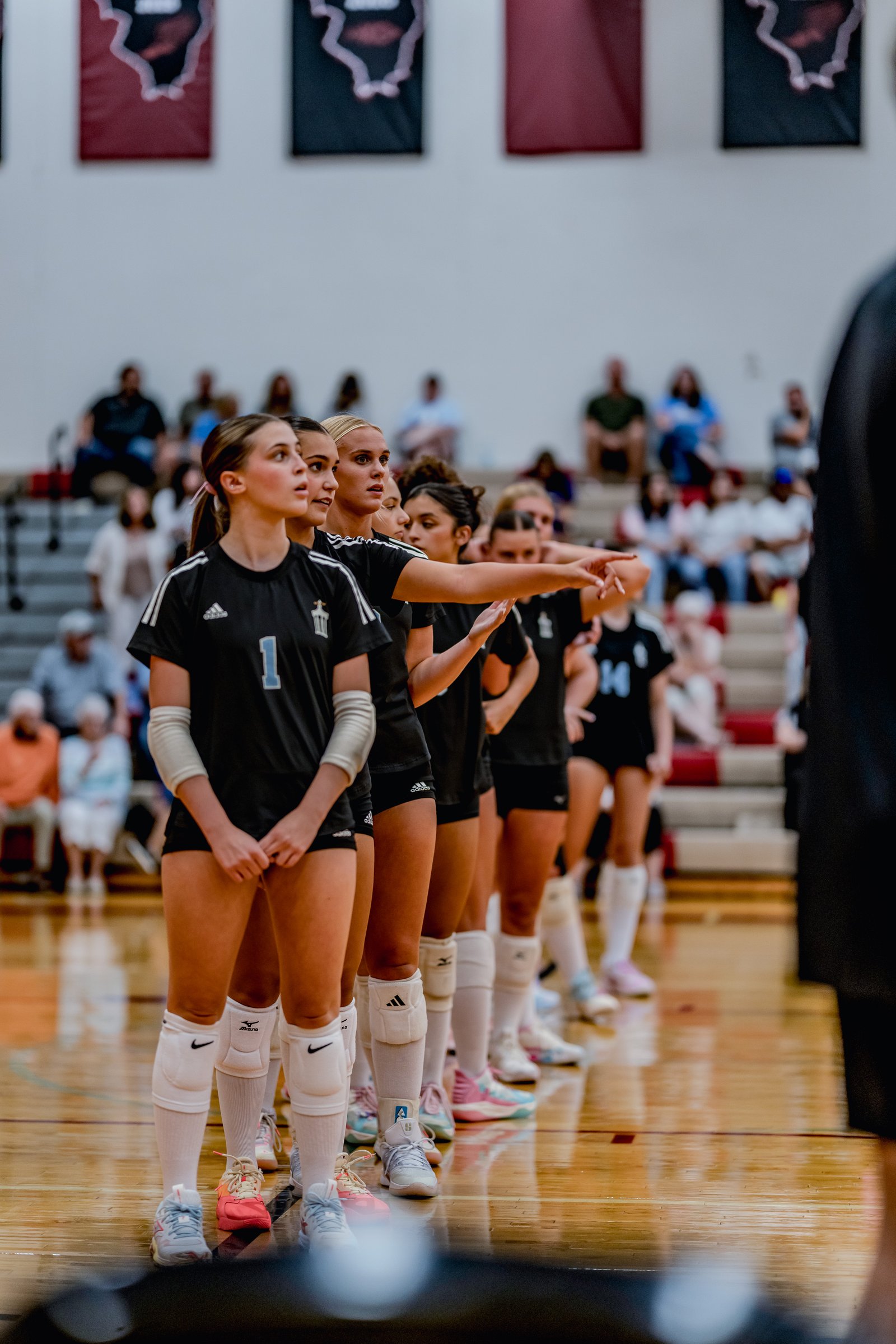 Players in black jerseys with white stripes stand on a wooden court, with one player pointing forward. Visible jersey number is 1.
