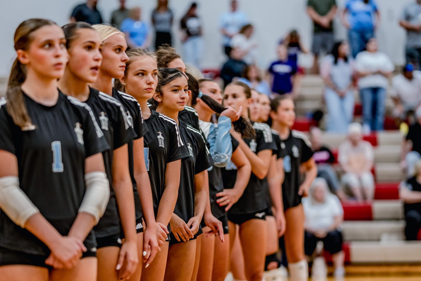 Players in black jerseys with white trim stand in a line on the court, with spectators in the background.