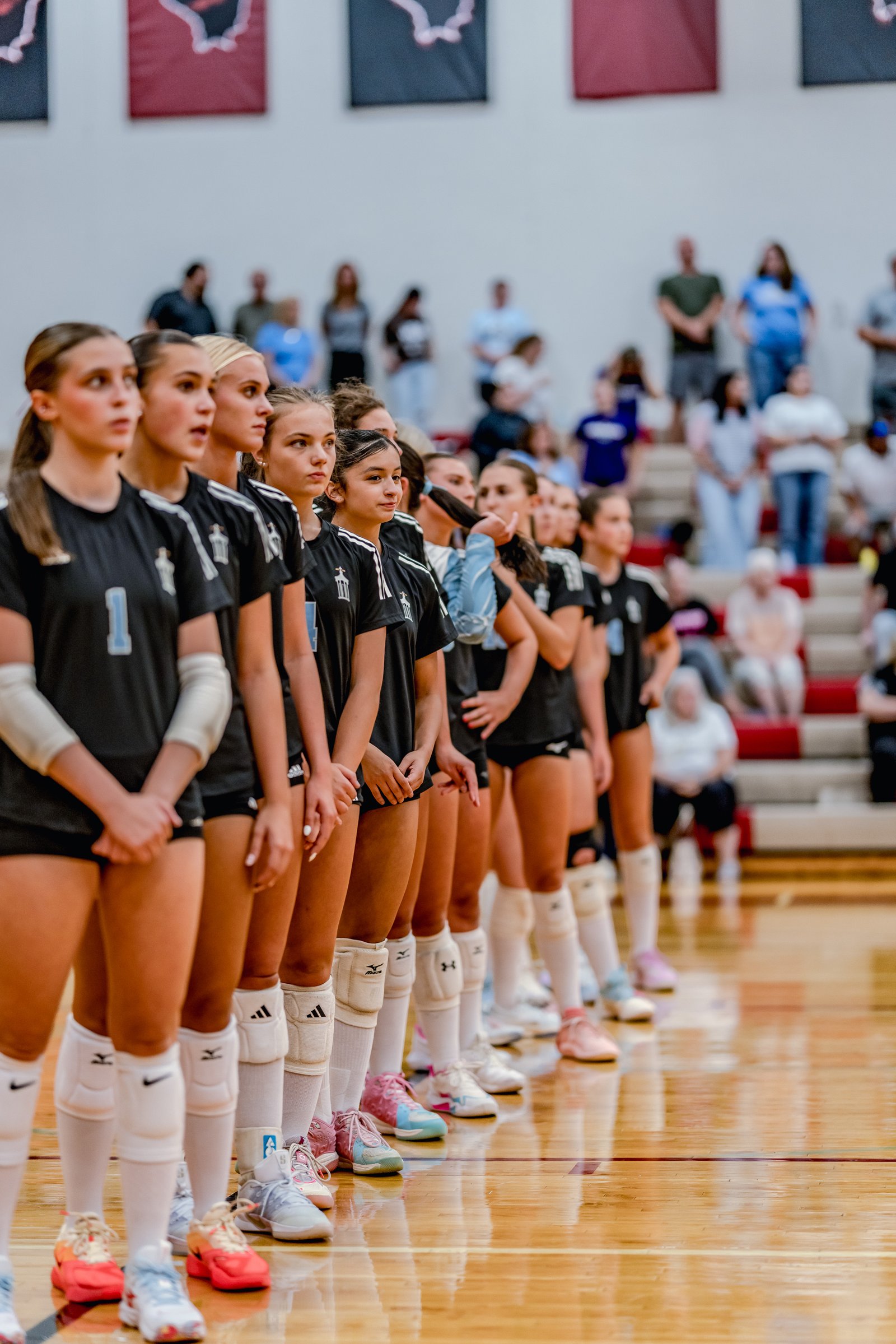 Players in black jerseys with white trim stand in a line on a wooden court, with spectators in the background.
