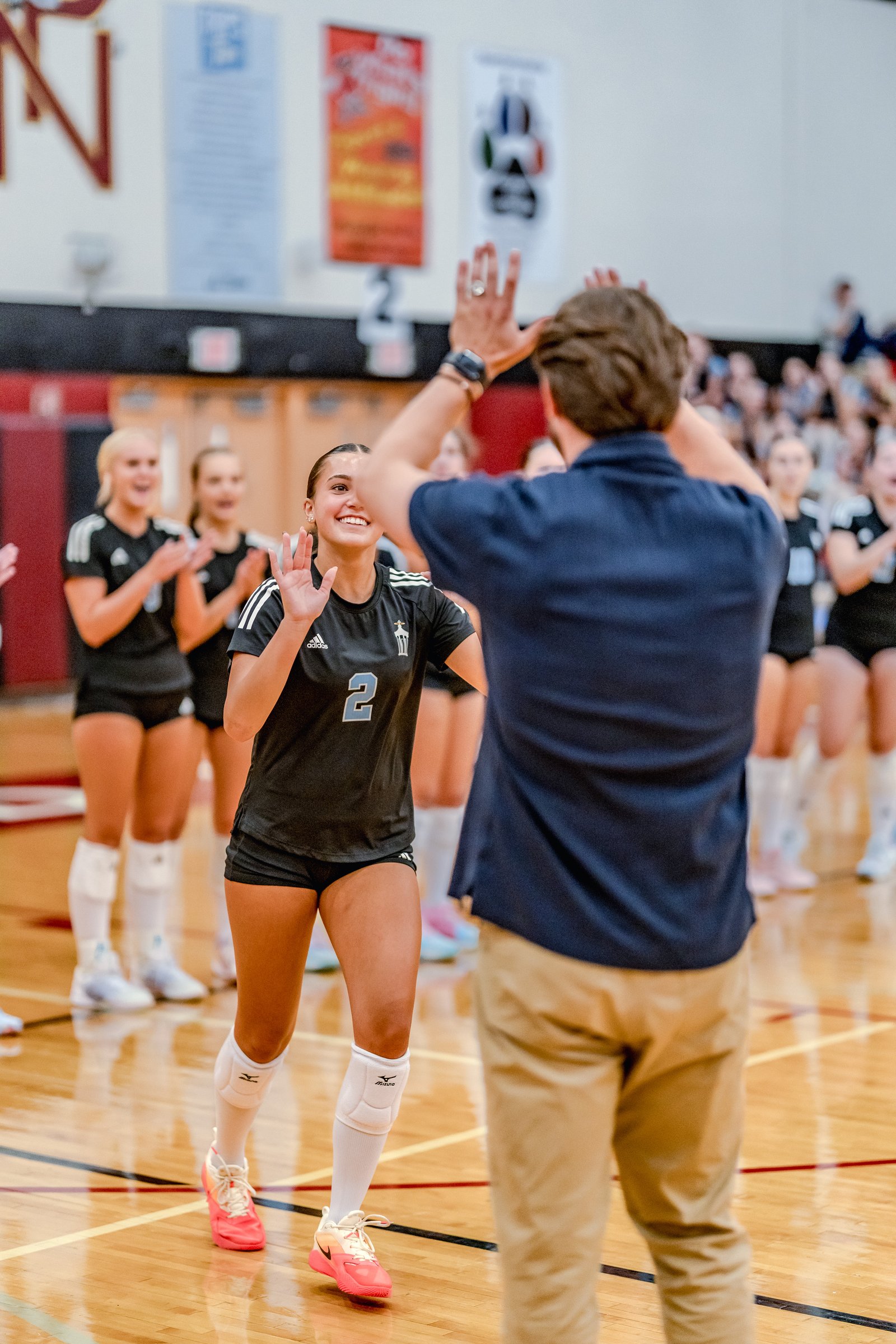 A player in black with jersey number 2 high-fives a coach on the court, with teammates in black and white in the background.