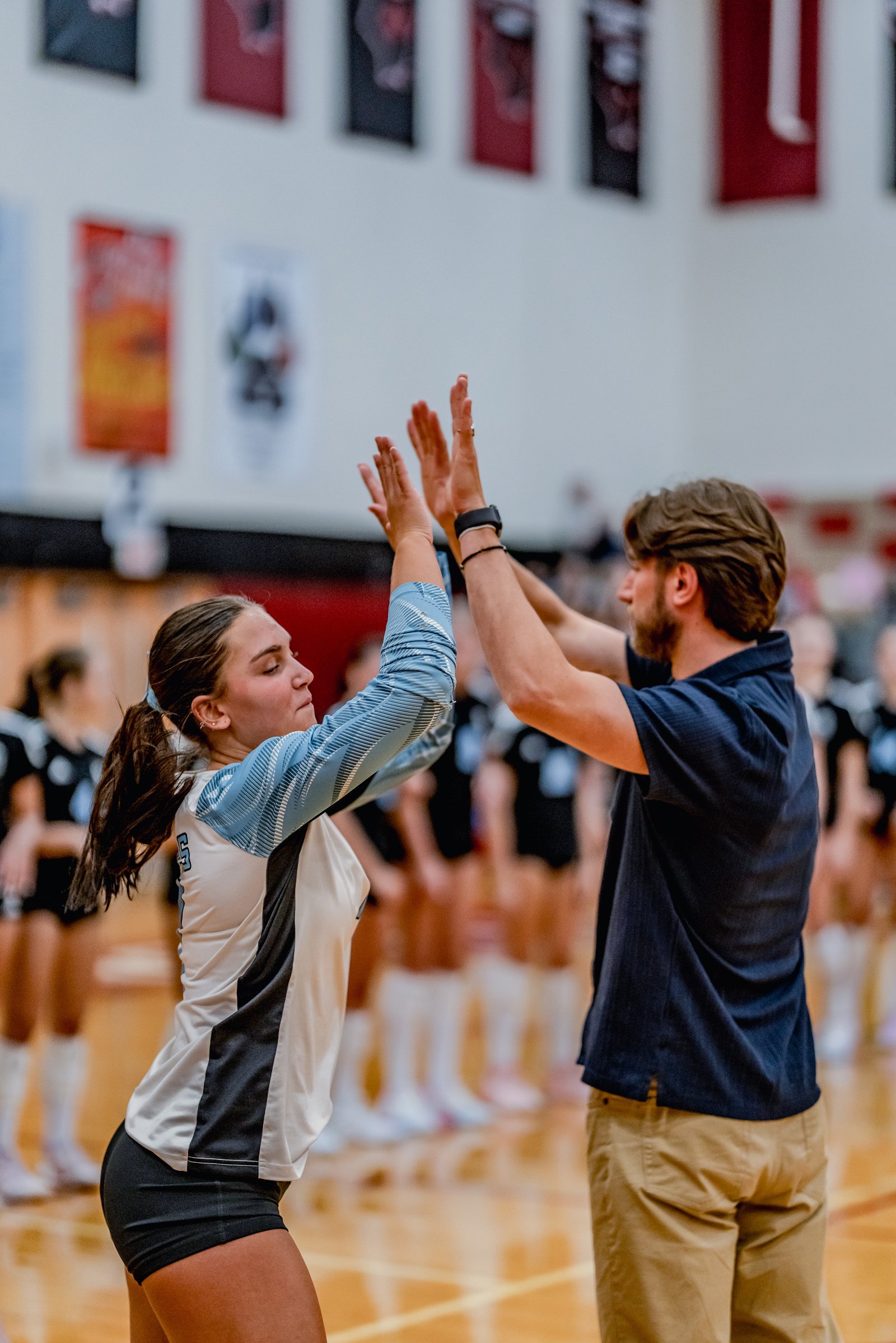 A player in a white and blue jersey high-fives a coach in a navy shirt and tan pants on the court.