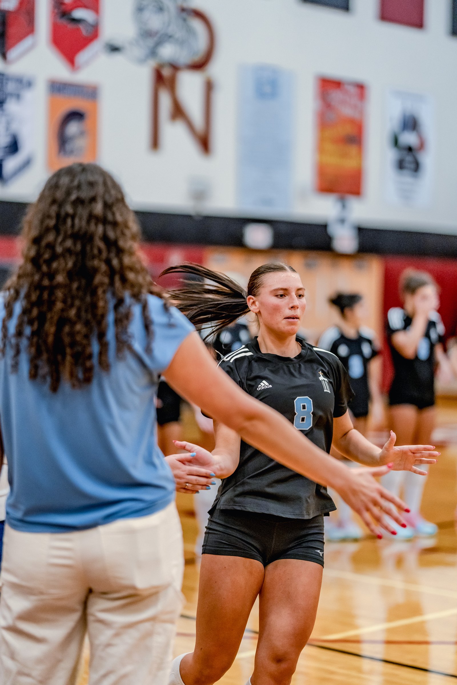 A player in a black jersey with blue number 8 and a player in a blue shirt interact on the court during a volleyball game.