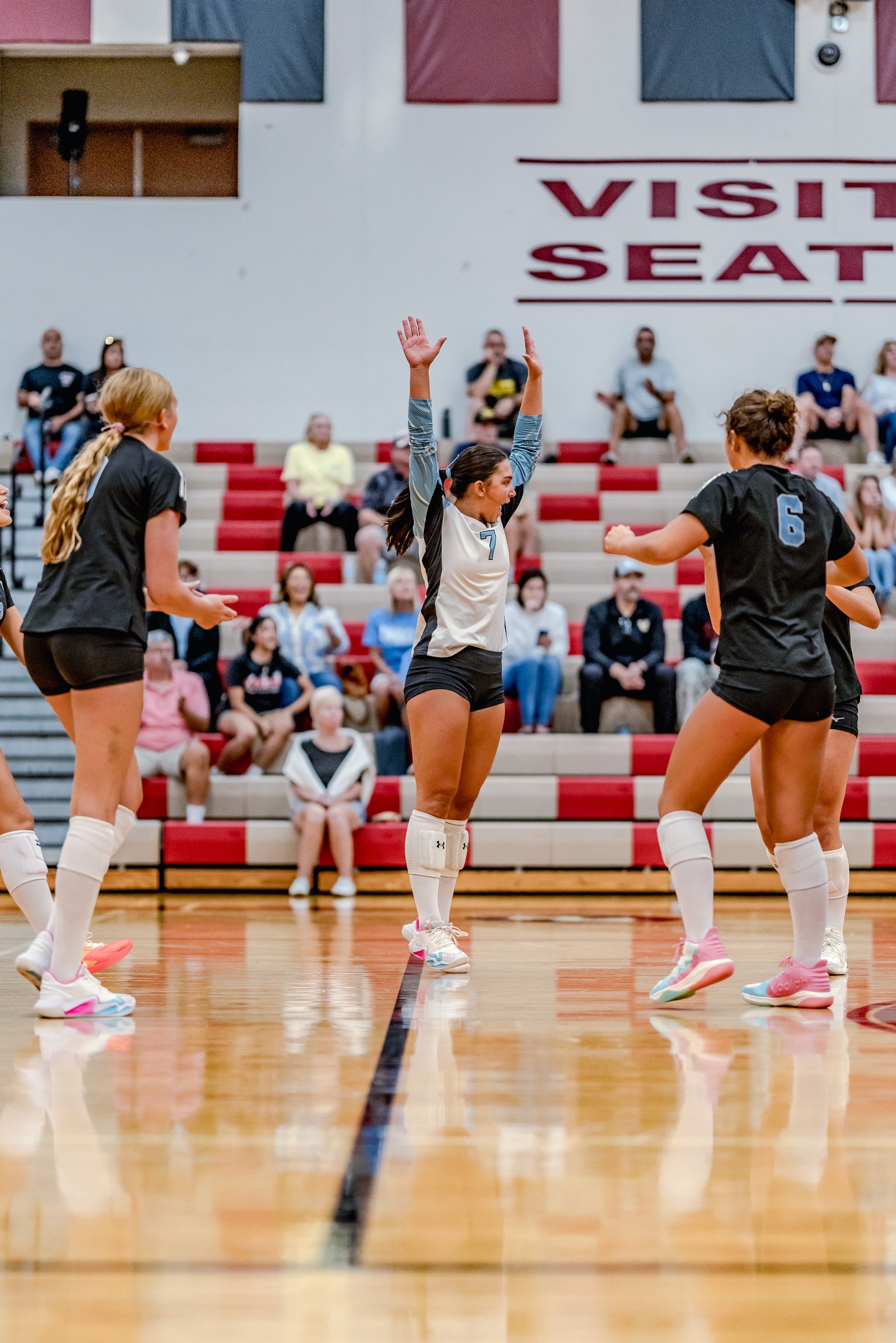 A player in a white and blue jersey with number 7 raises her arms, while players in black jerseys with numbers 6 and 00 are visible on the court.