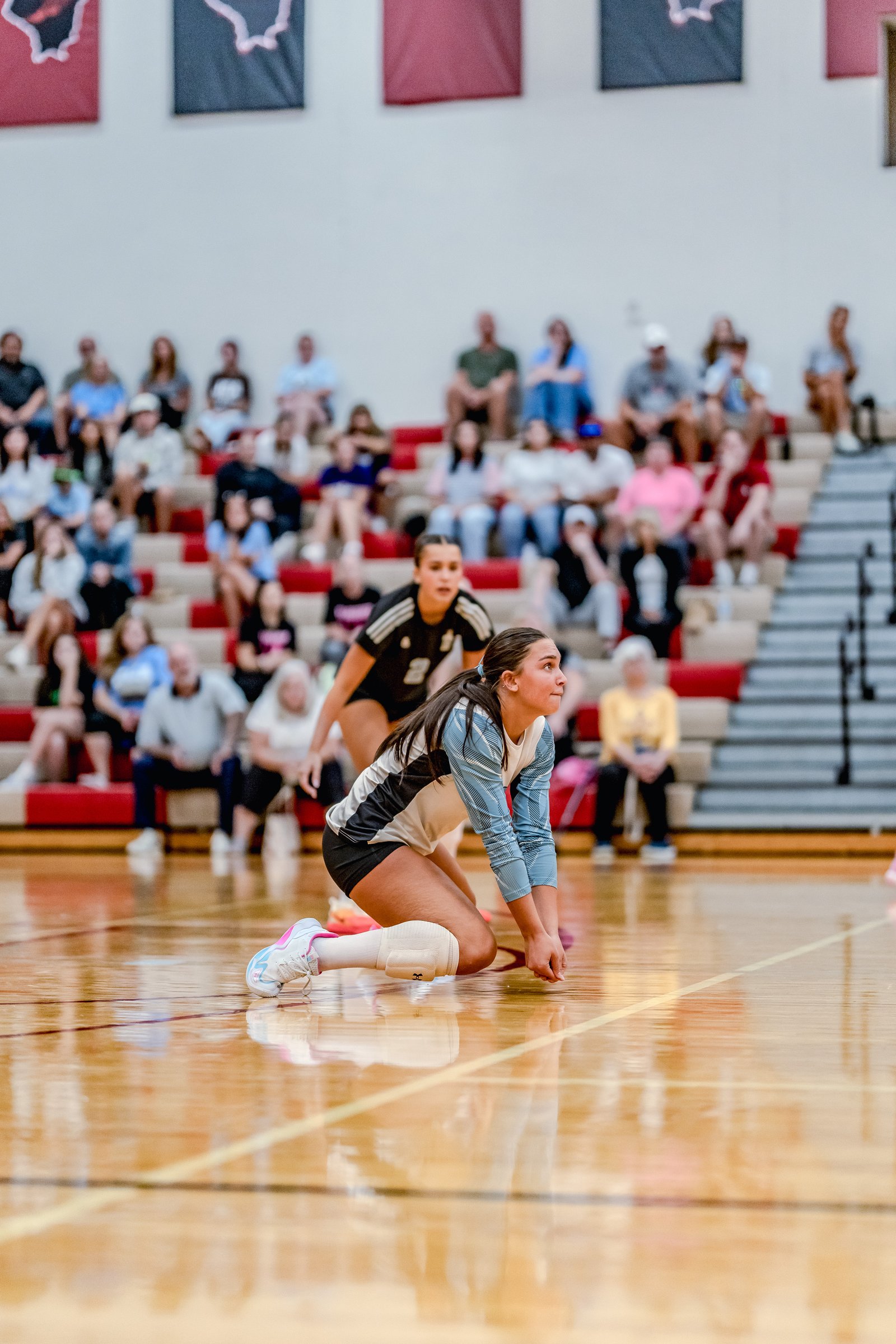 A player in a light blue and white jersey prepares to dig a volleyball on the court, with spectators in the background.