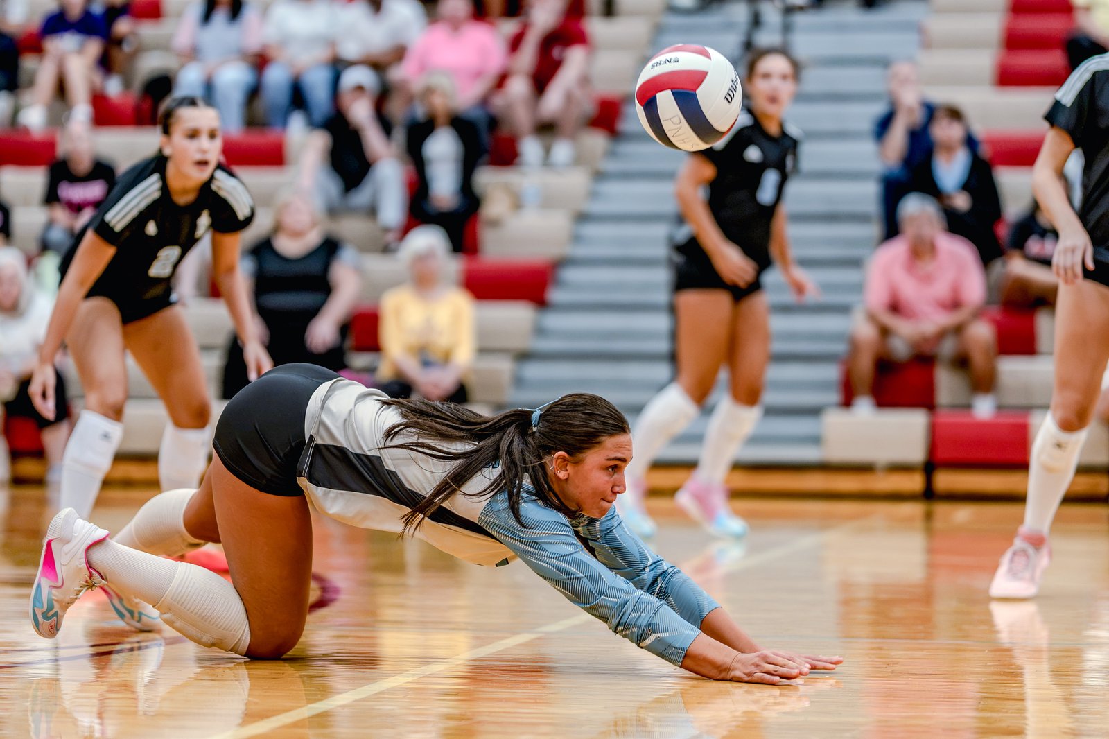 A player in a light blue and white jersey dives for a volleyball, with players in black jerseys visible in the background.
