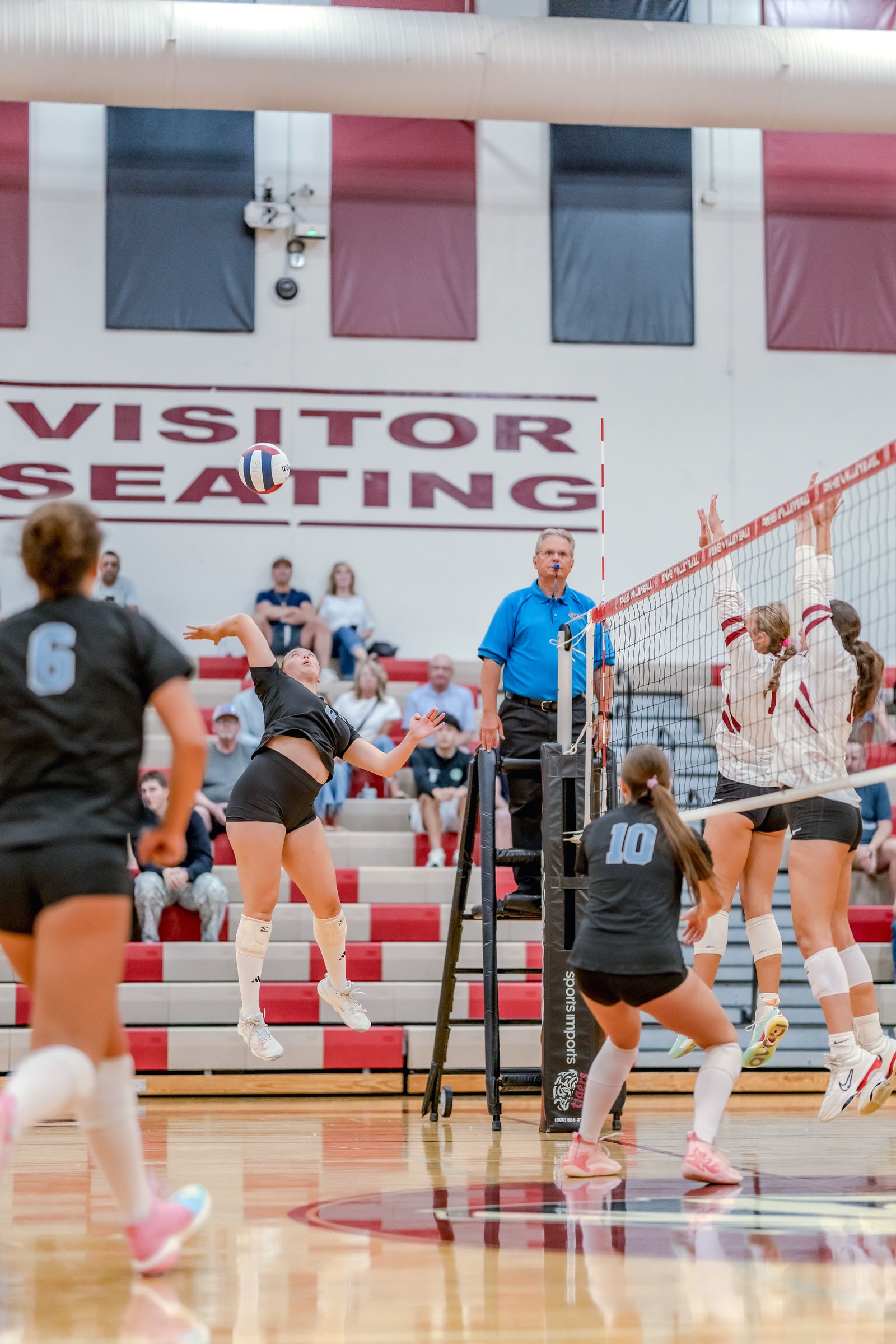 A player in black jumps to hit a volleyball while players in white and maroon attempt to block, with a referee watching.
