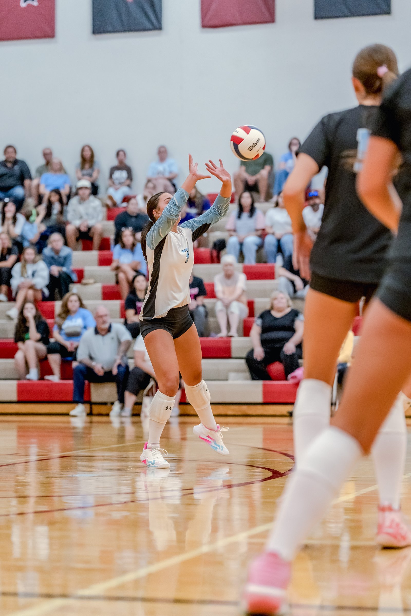 A player in a white and blue jersey with number 7 sets the volleyball during a game with spectators in the background.