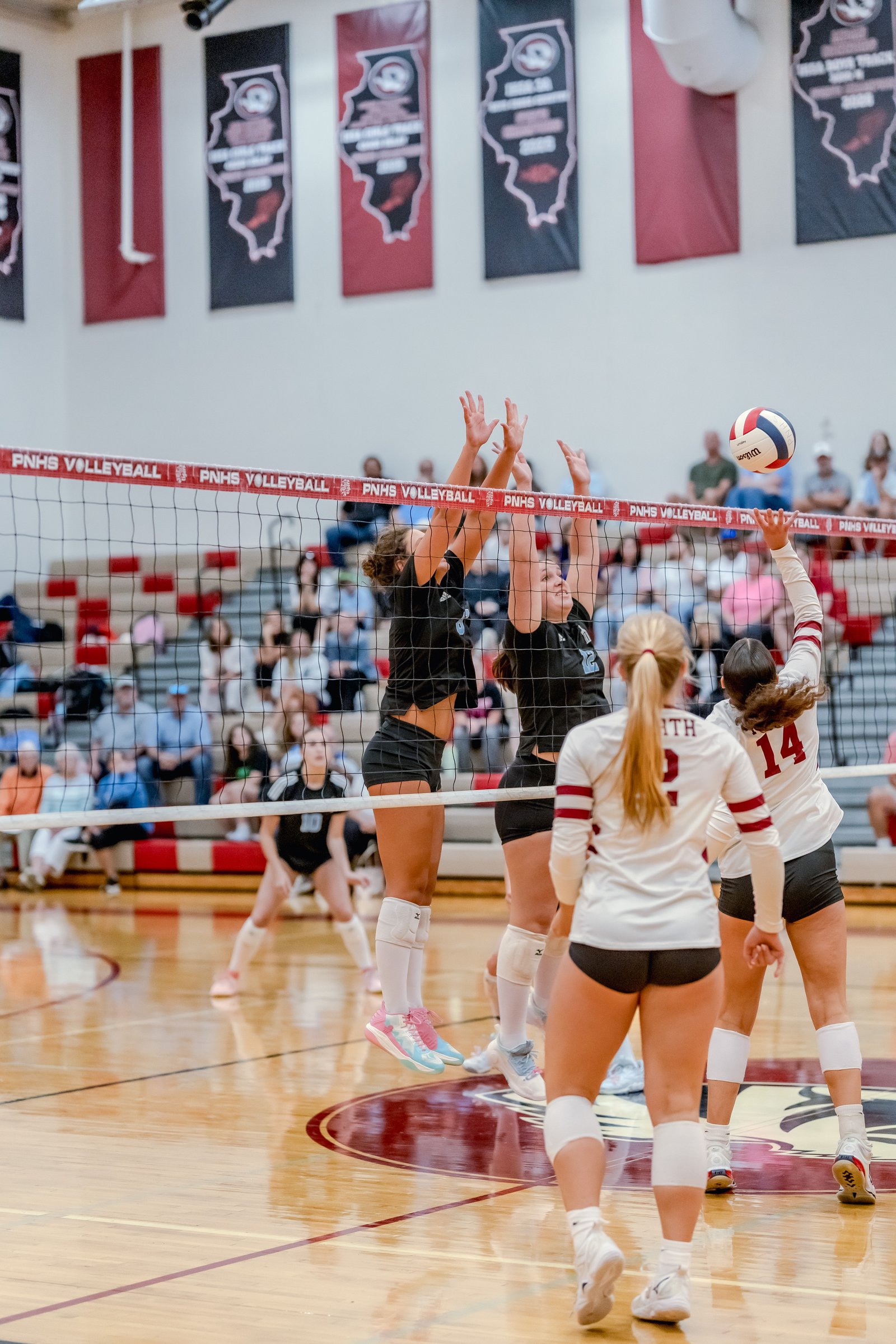 Players in black and white jerseys jump at the net during a volleyball game, with a ball in the air.