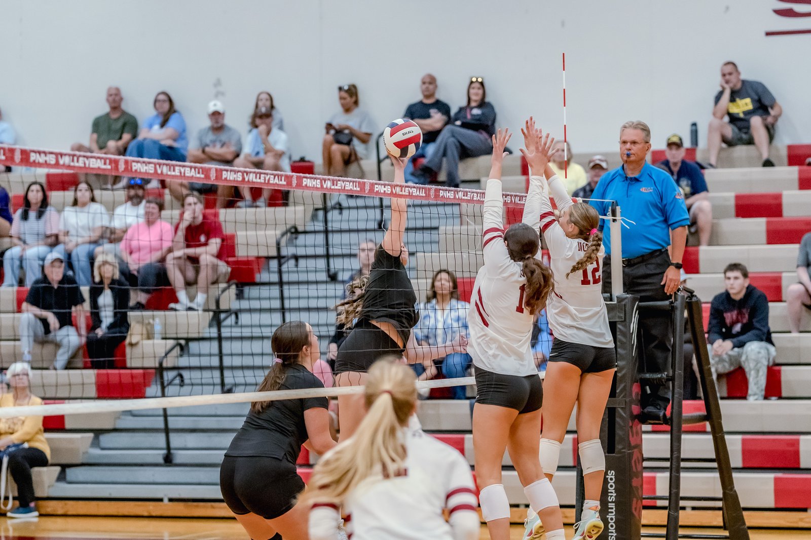 A player in black spikes the ball over the net as players in white attempt to block during a volleyball game.