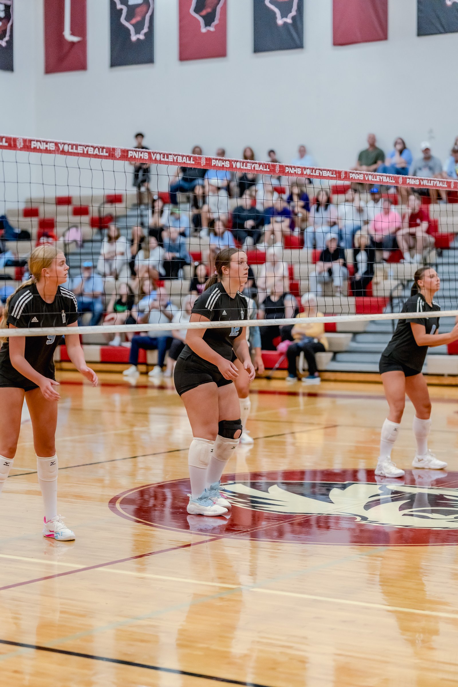 Three players in black tops and shorts are on the court during a volleyball game, with spectators in the background.