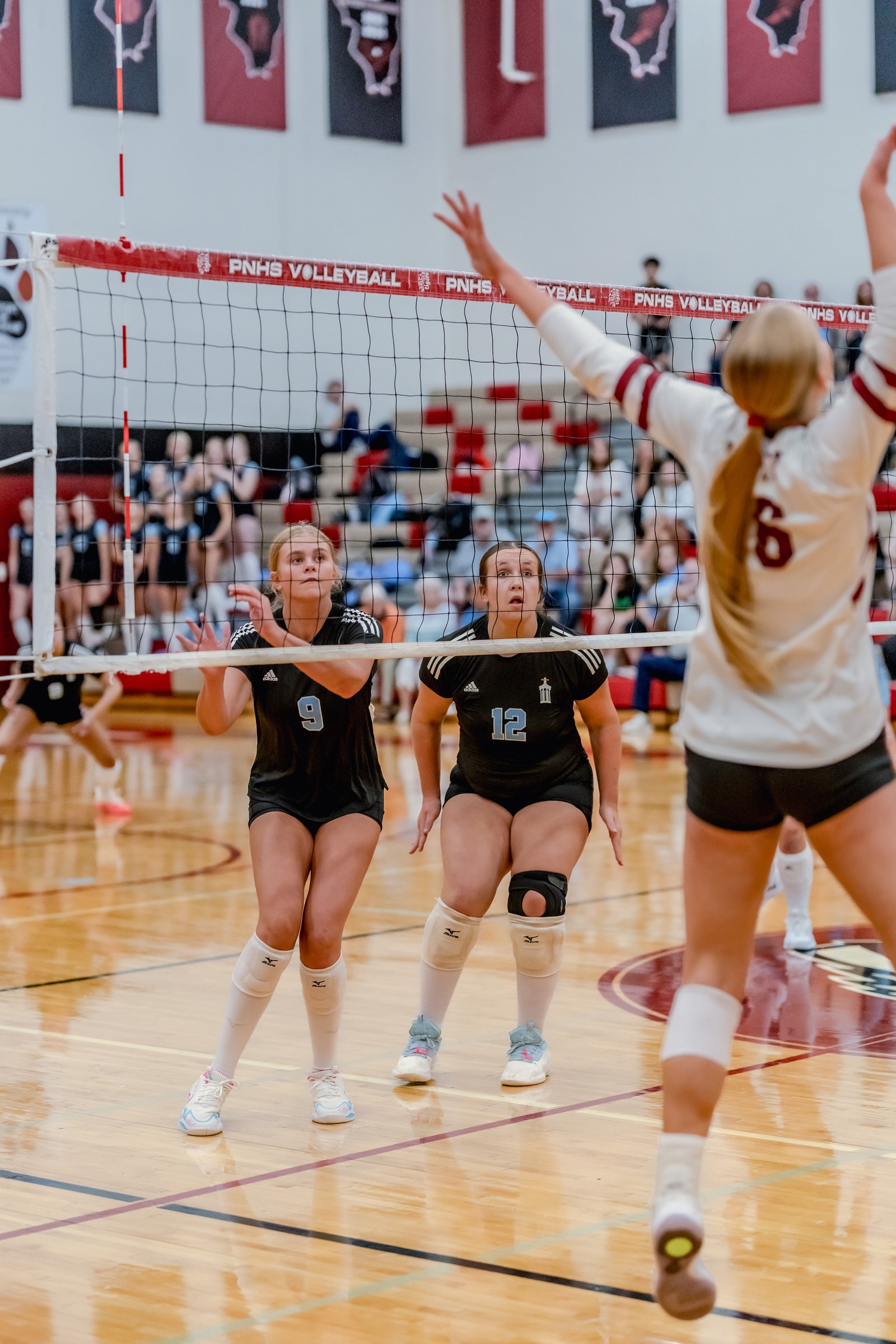 Players in black and white uniforms prepare for a volleyball play near the net, with one player in a white uniform jumping.