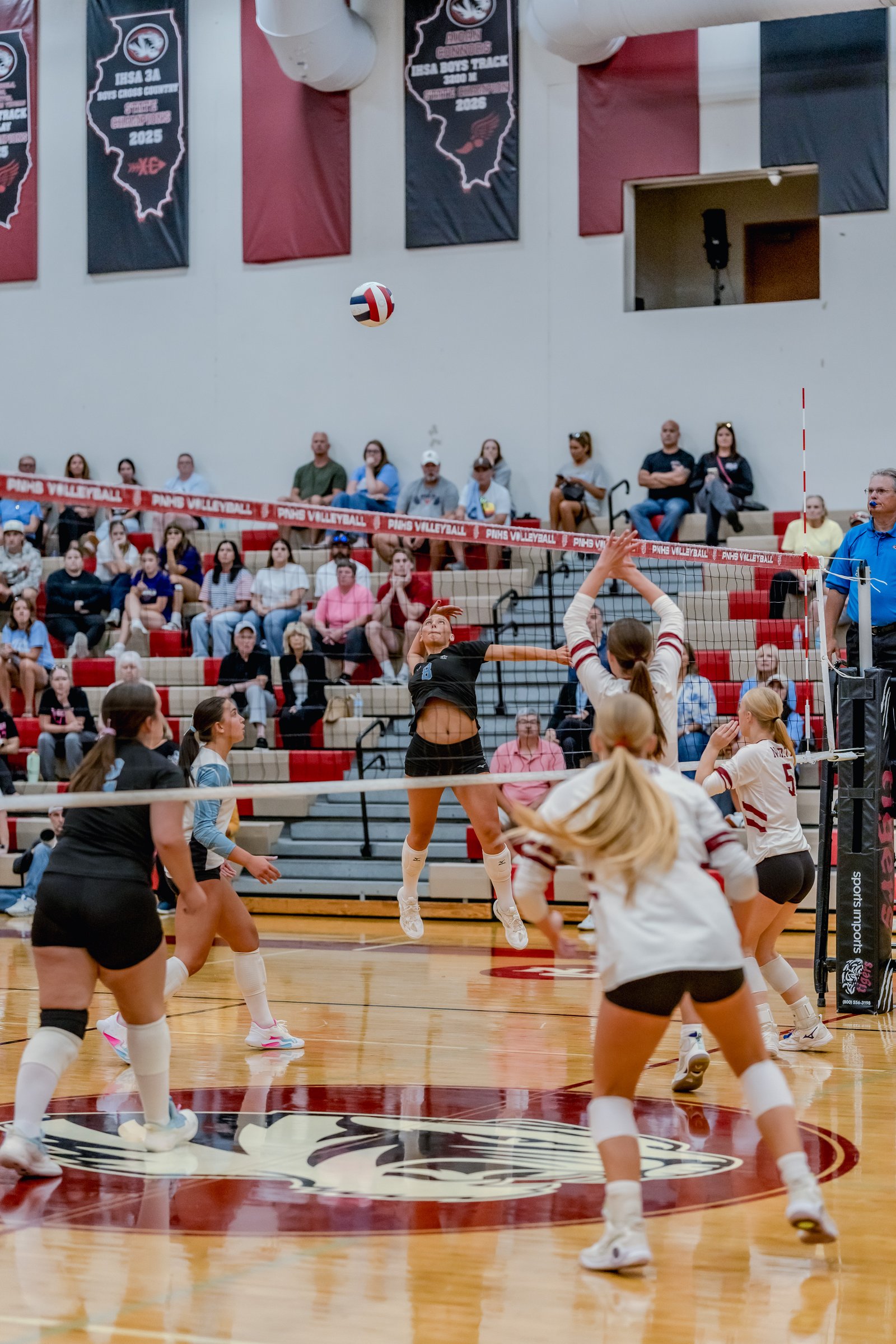 A player in black jumps to spike a volleyball during a game, with players in white and maroon on the court and spectators in the stands.