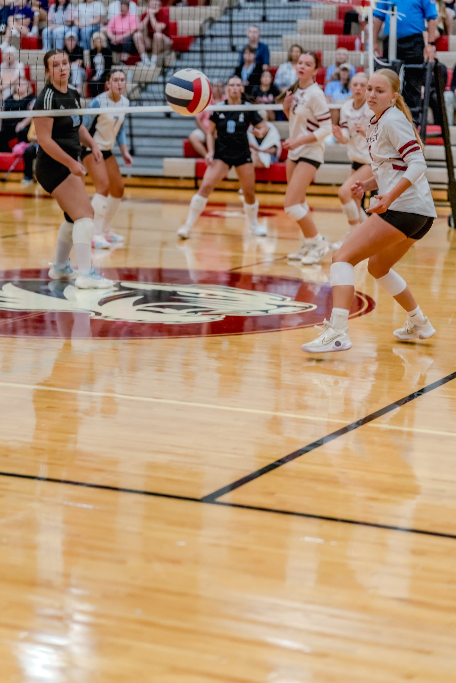 A player in white with maroon trim passes a volleyball over the net while teammates in white and a player in black watch.