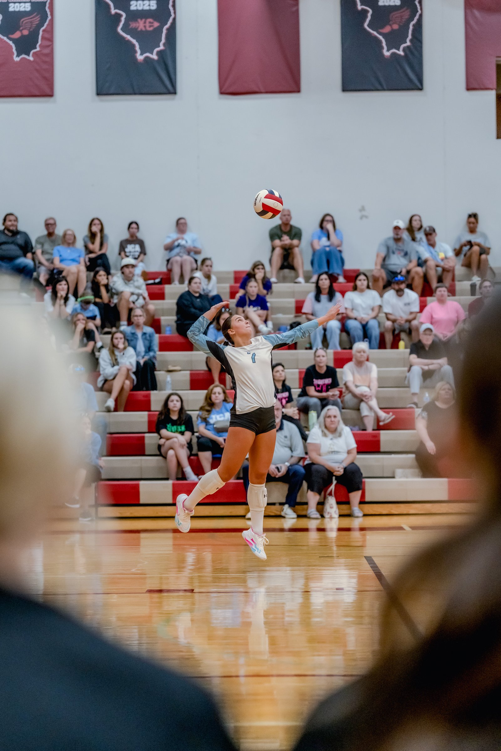 A player in a light blue and white jersey serves a volleyball during a game in a gymnasium with spectators in the background.