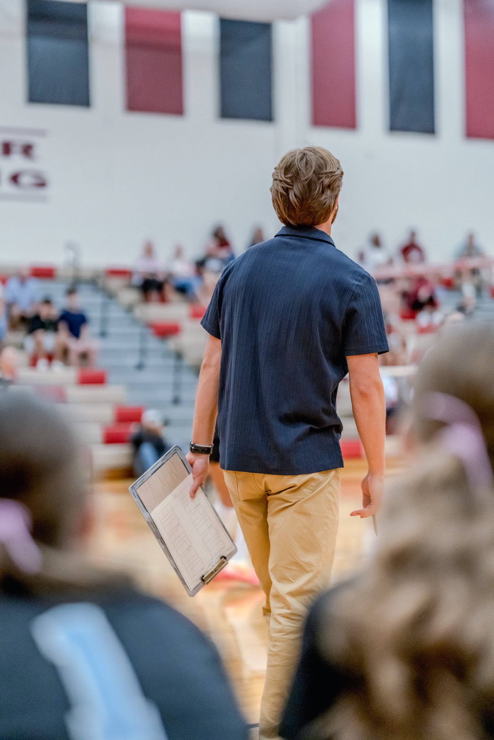 A coach in a navy shirt and tan pants walks with a clipboard on the court, with blurred players and spectators in the background.