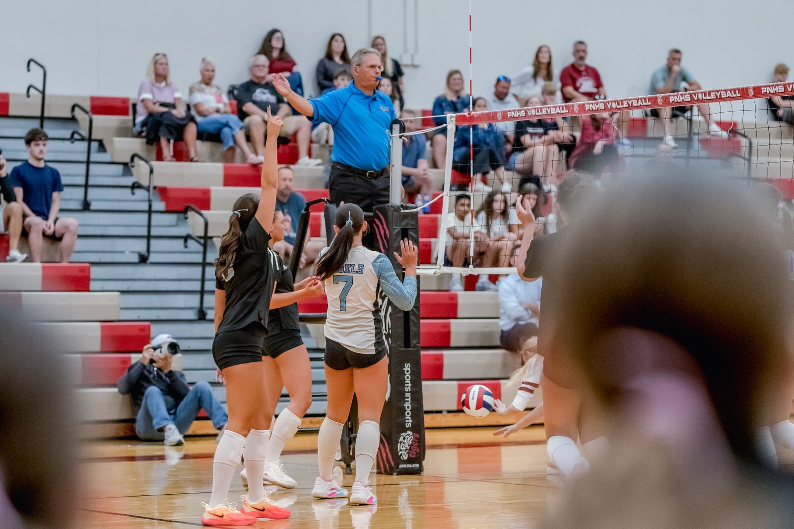 Players in black and white jerseys compete in a volleyball game, with a referee signaling and spectators in the background.
