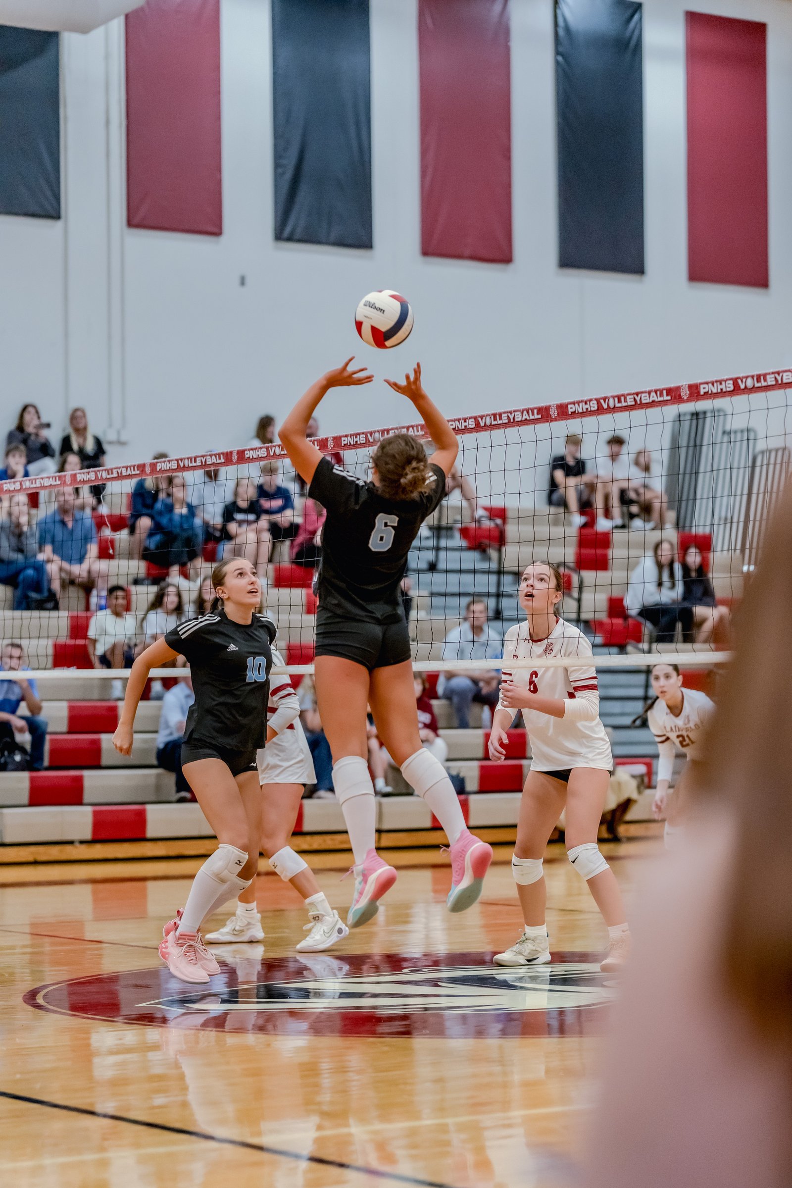 A player in black with jersey number 6 sets the volleyball as players in black with number 10 and white with number 6 look on.