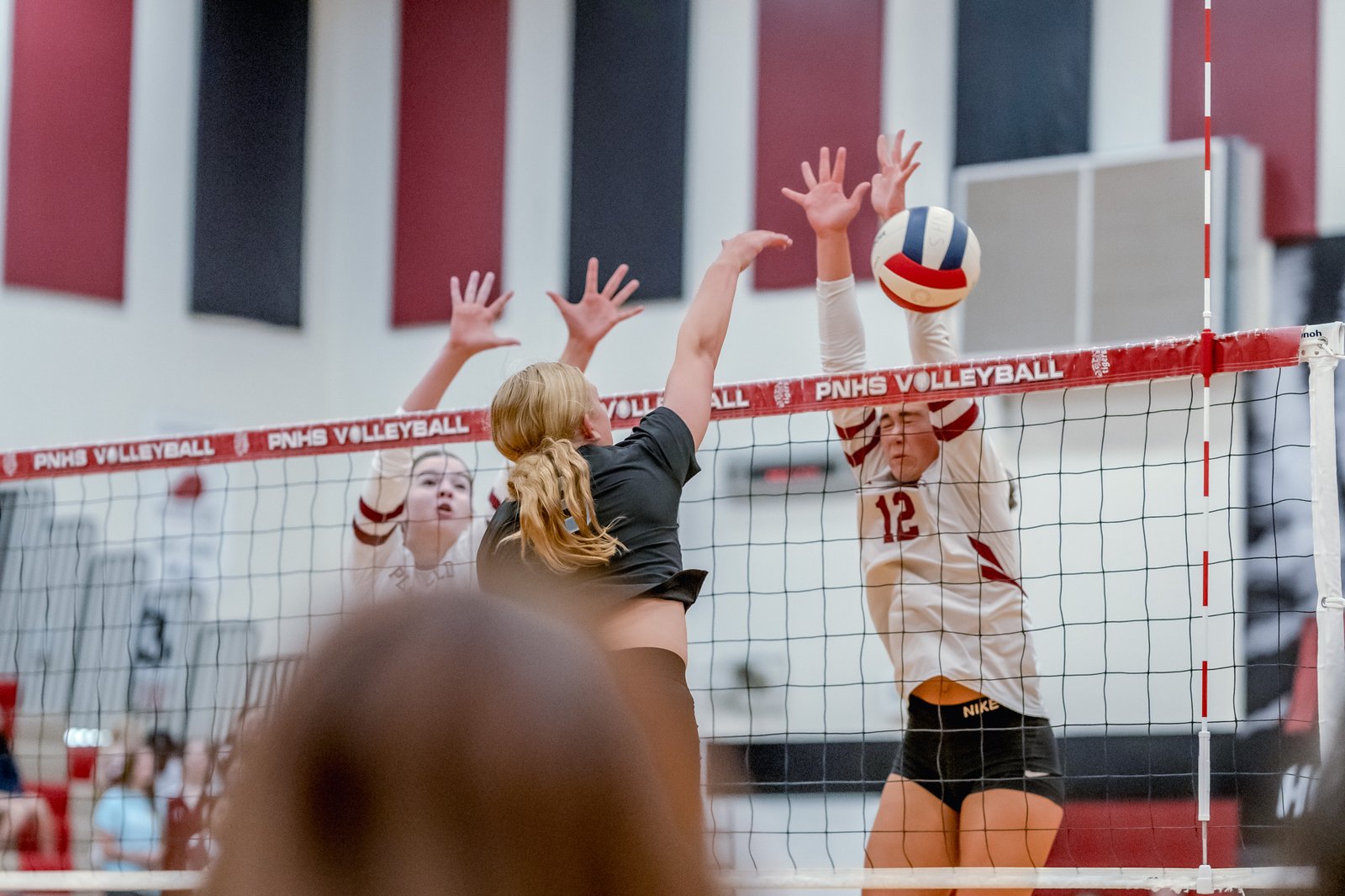 A player in a black top attempts a spike over the net as players in white jerseys with red trim and number 12 jump to block.
