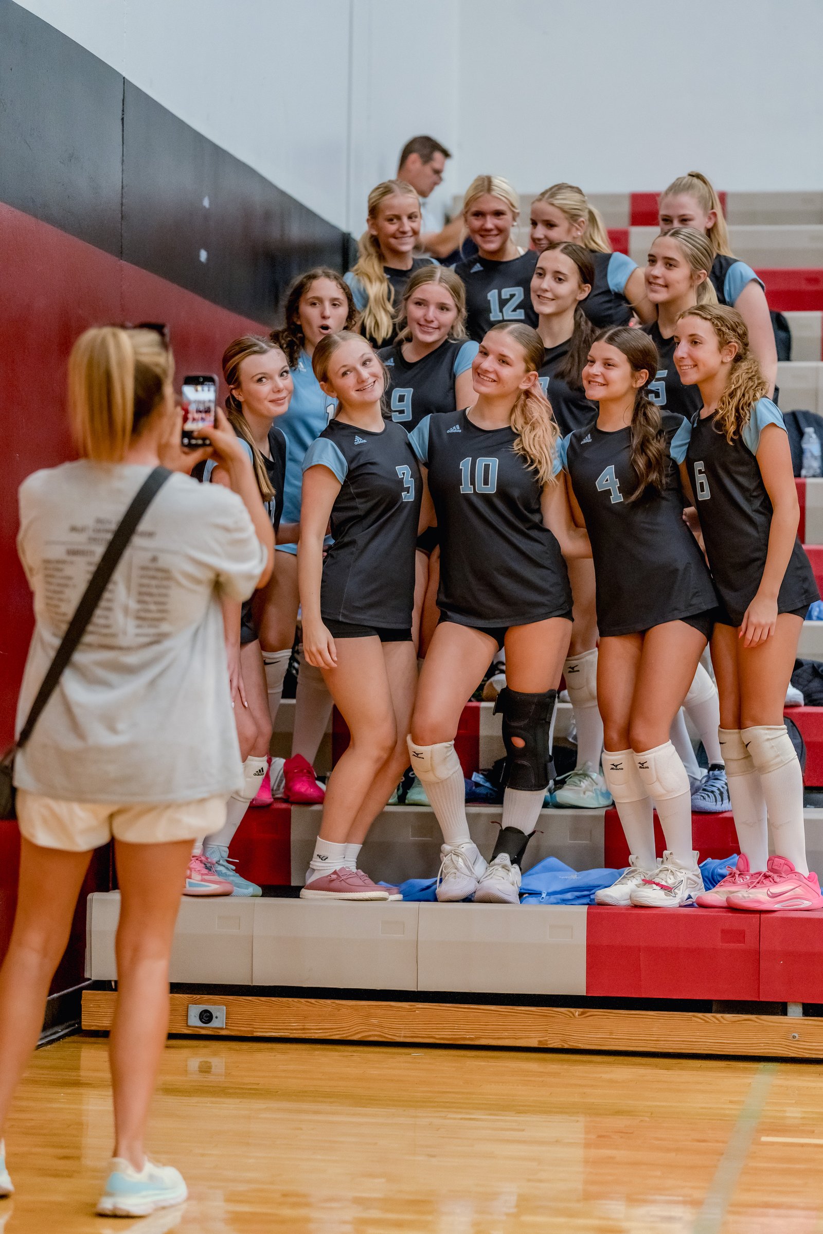A group of volleyball players in black and light blue jerseys pose for a photo on the bleachers, with one player taking a picture with her phone.