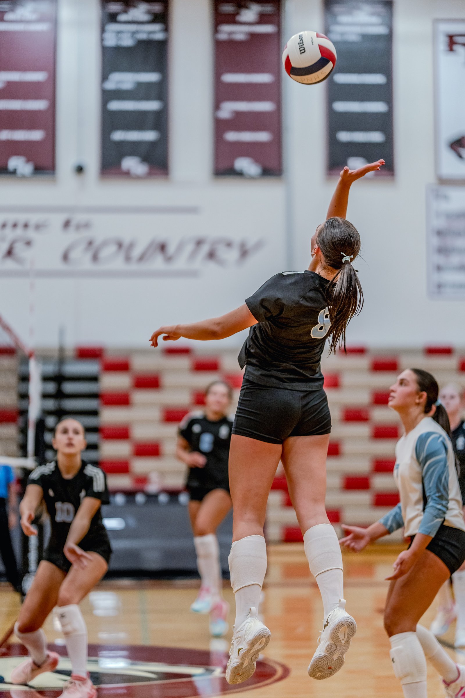 A player in black serves a volleyball over the net while players in black and light blue watch on the court.