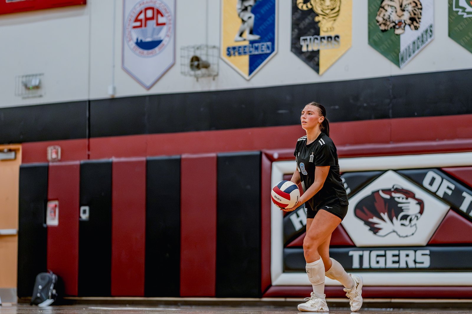 A player in a black top with white stripes holds a volleyball, preparing to serve on a court with team banners.