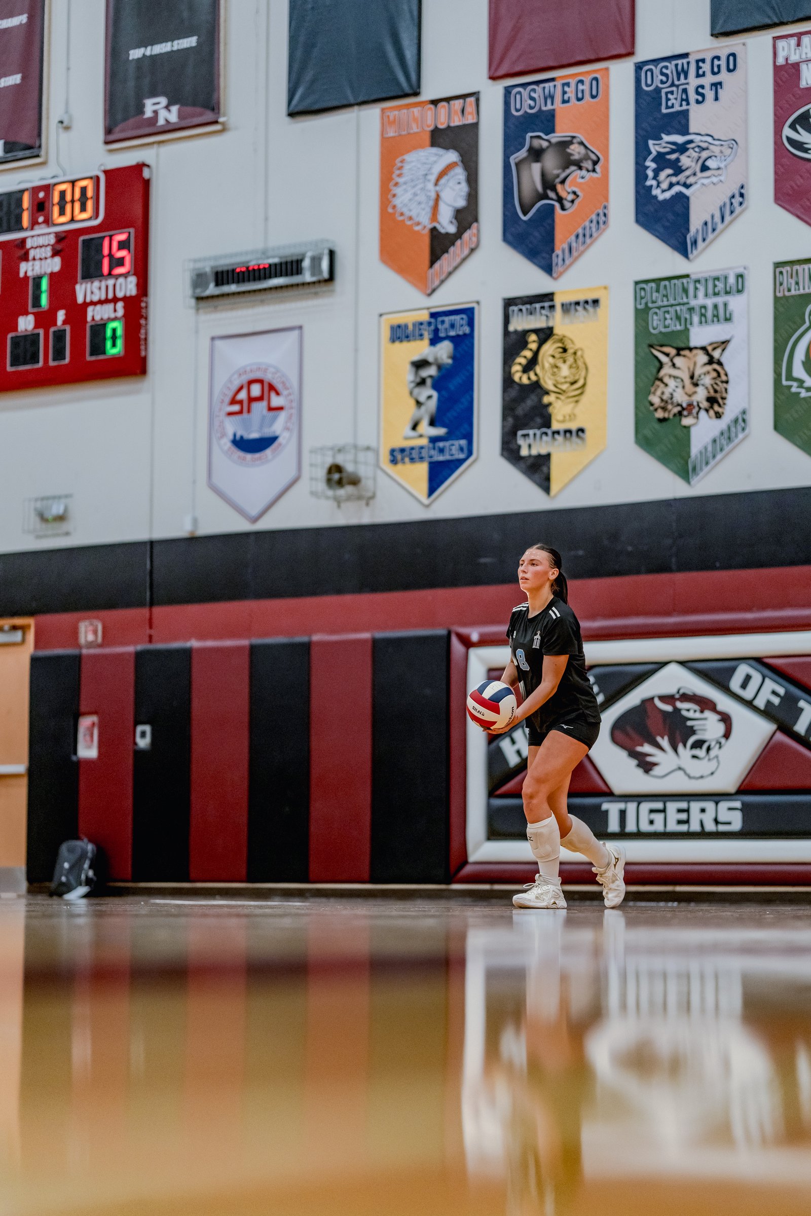 A player in a black top and shorts prepares to serve a volleyball in a gymnasium with banners on the wall.