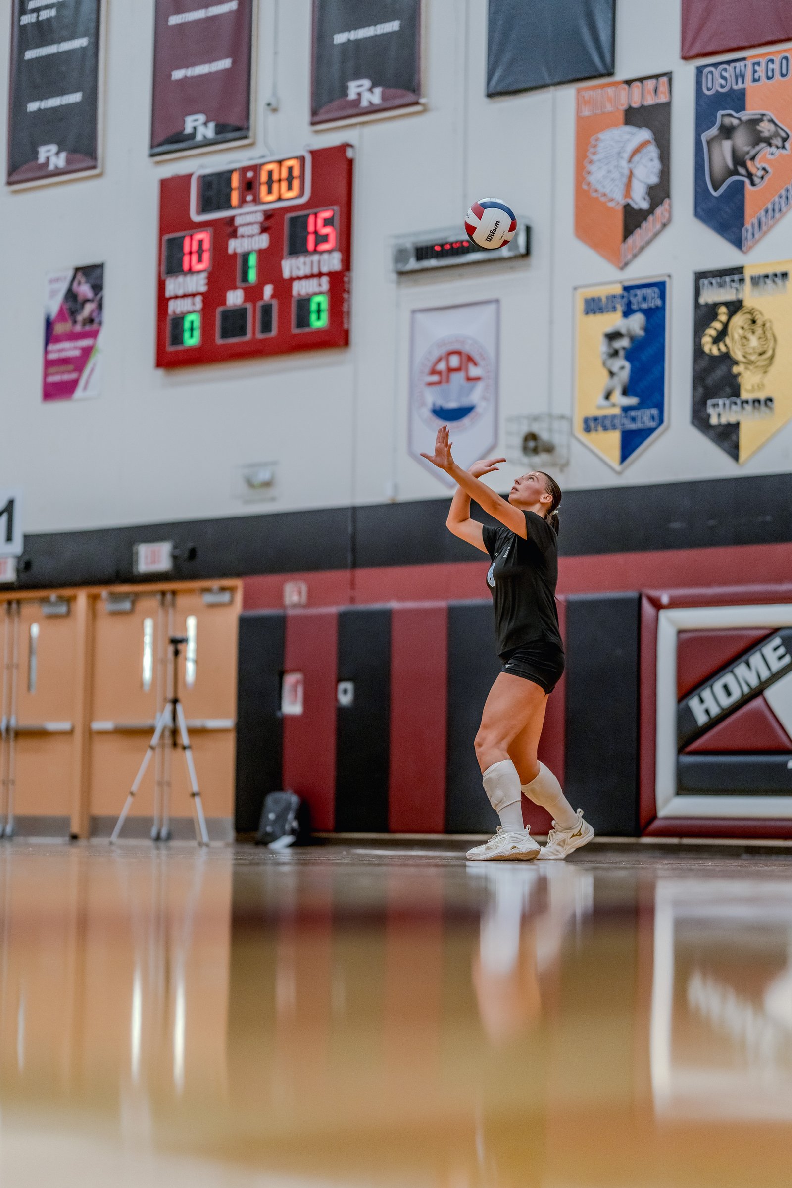 A player in a black top sets the volleyball during a game, with a scoreboard and team banners in the background.