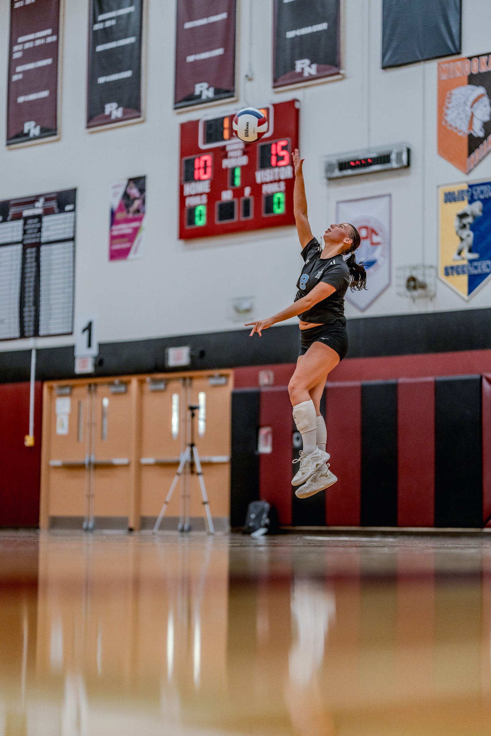 A player in a black top and shorts jumps to hit a volleyball during a game, with a scoreboard in the background.