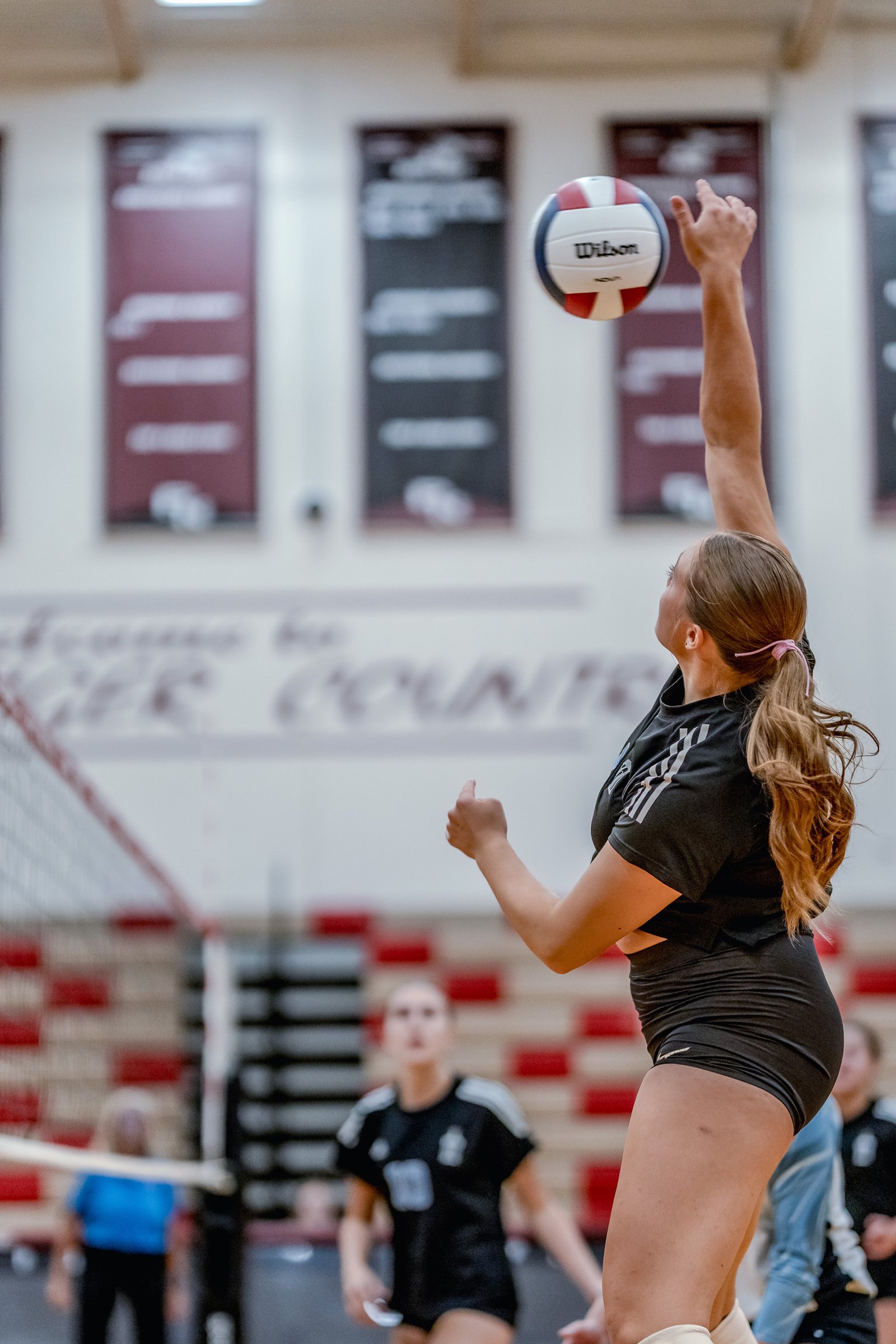 A player in a black top serves a volleyball during a game, with other players visible in the background.