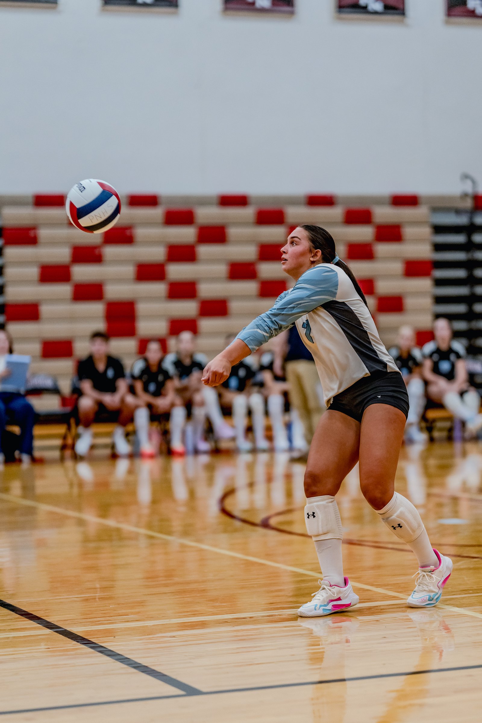 A player in a white and blue jersey passes a volleyball during a game. Other players sit on the bench in the background.