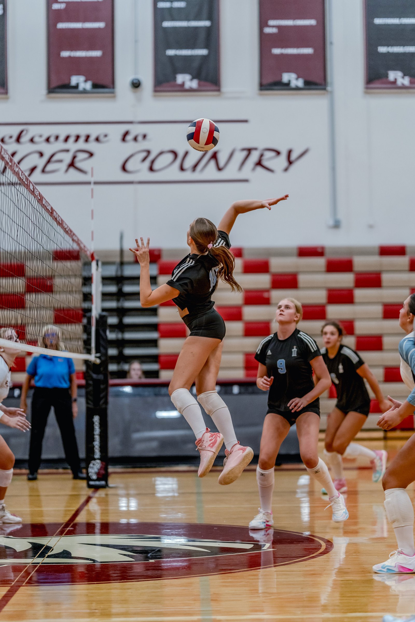 A player in black jumps to hit the volleyball over the net, with players in black and light blue watching.