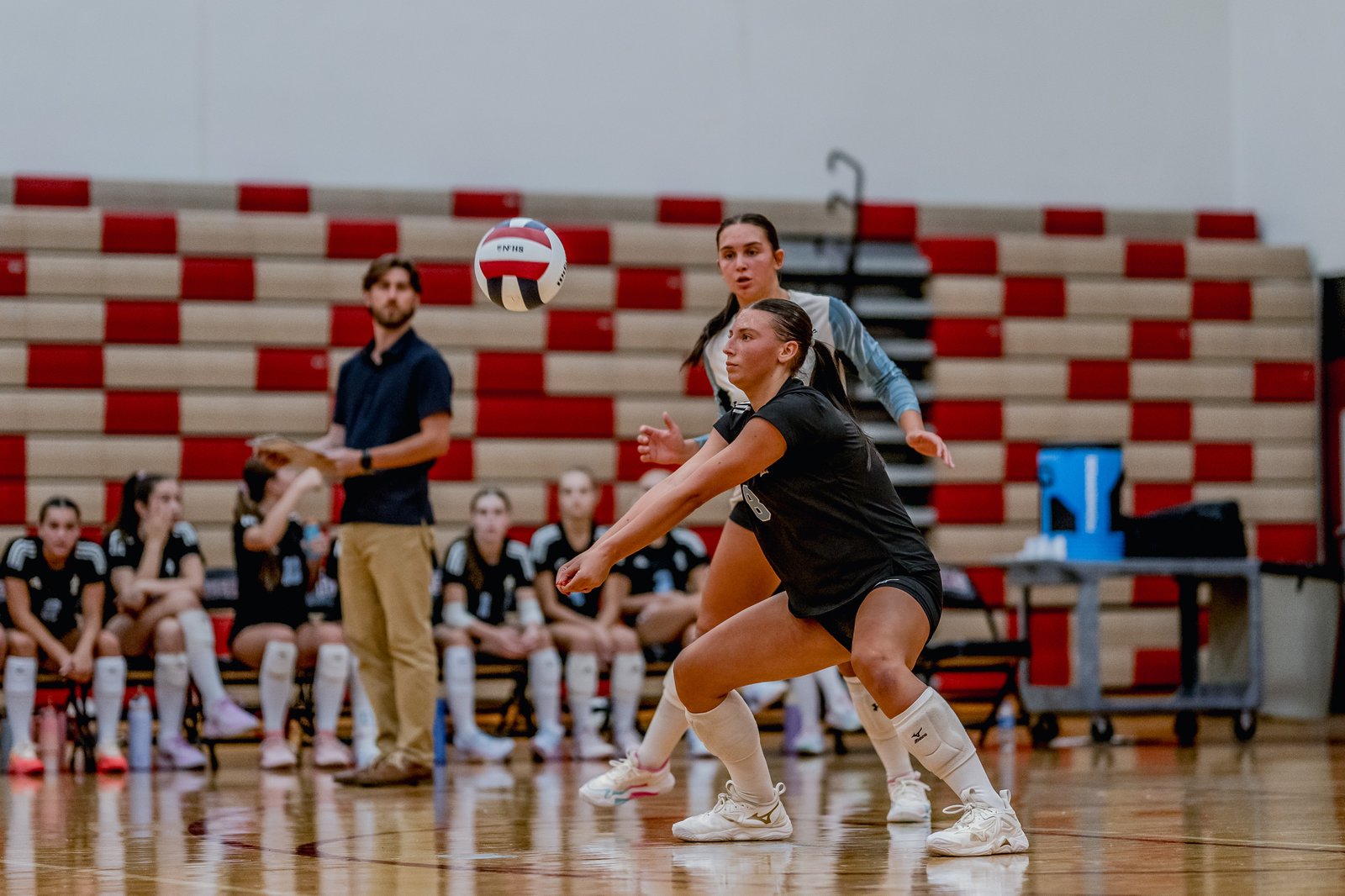 A player in black attempts a dig as a player in light blue watches the ball in a gymnasium with red and white bleachers.