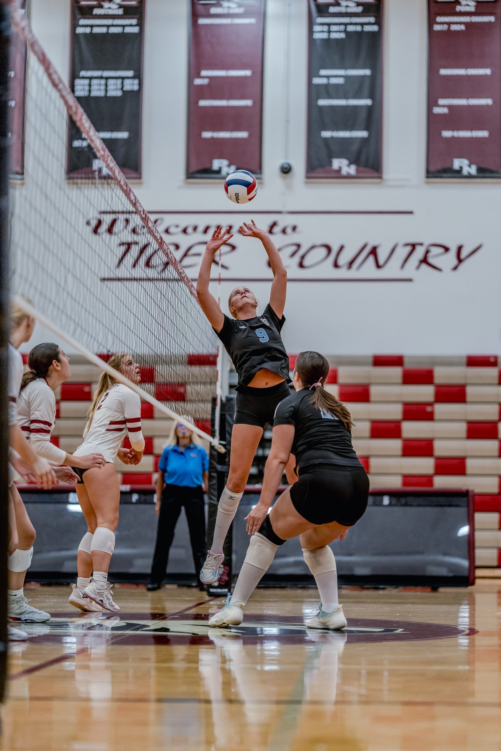 A player in black with jersey number 9 sets the volleyball during a game against players in white and maroon.