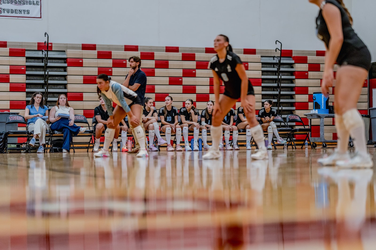 A player in a black top and shorts prepares to play, while teammates in black and white sit on the bench in a gymnasium.