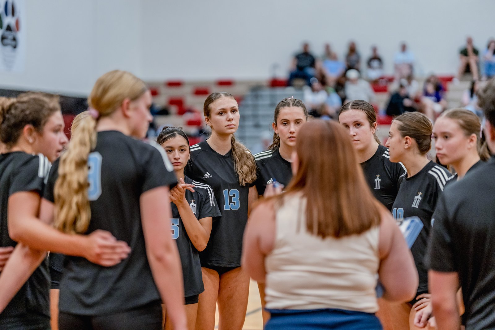 Players in black jerseys with blue numbers huddle together on the court, listening intently.