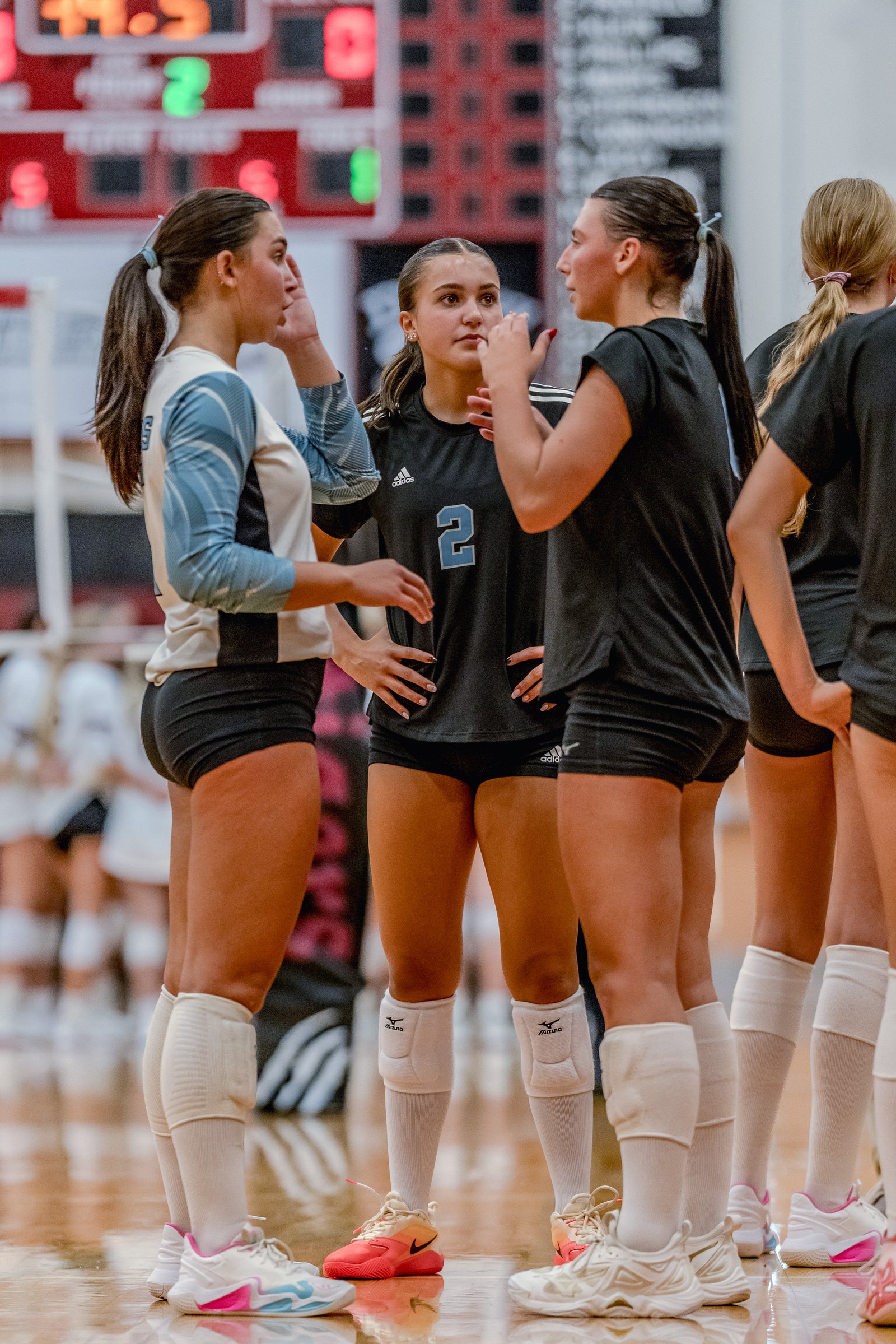 Players in black and light blue jerseys huddle on the court, with a scoreboard in the background.