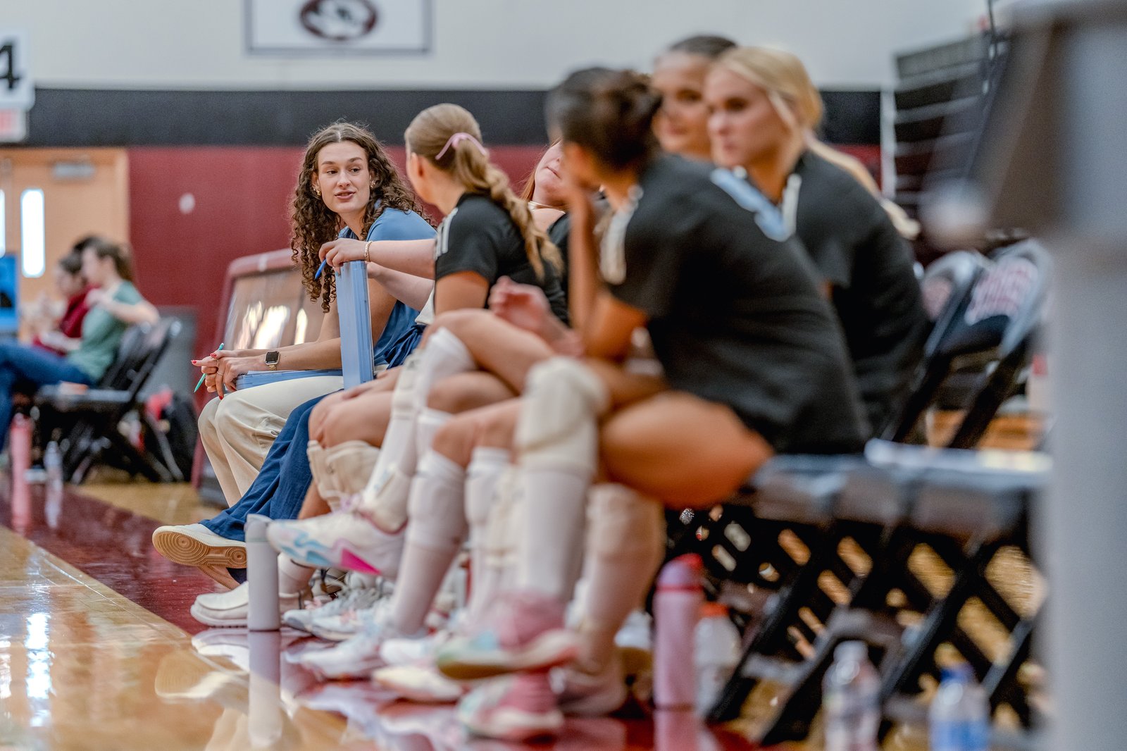 A player in a blue top talks to teammates in black tops on the bench during a volleyball game.