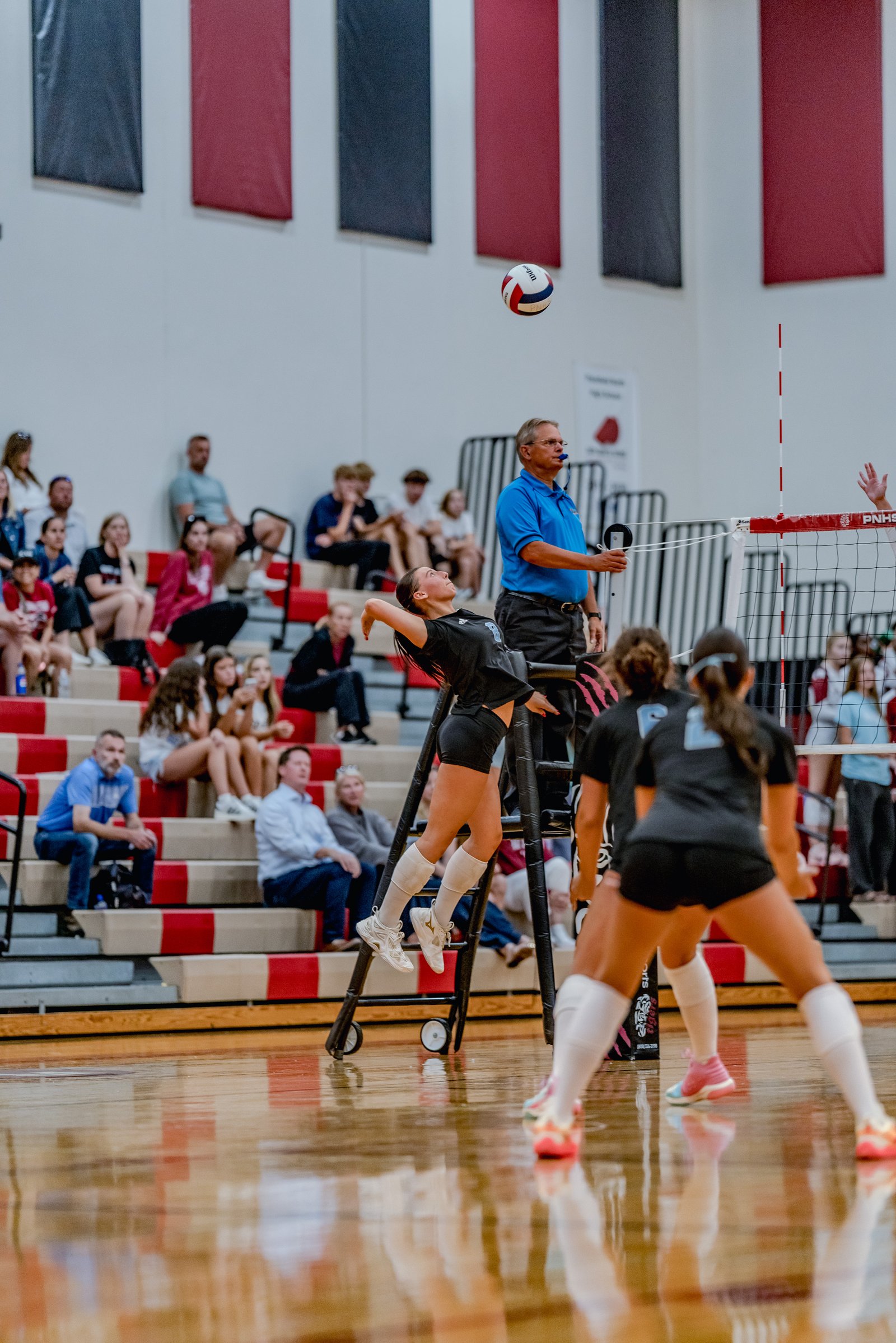 A player in a black top jumps to hit the volleyball during a game, with spectators in the background.