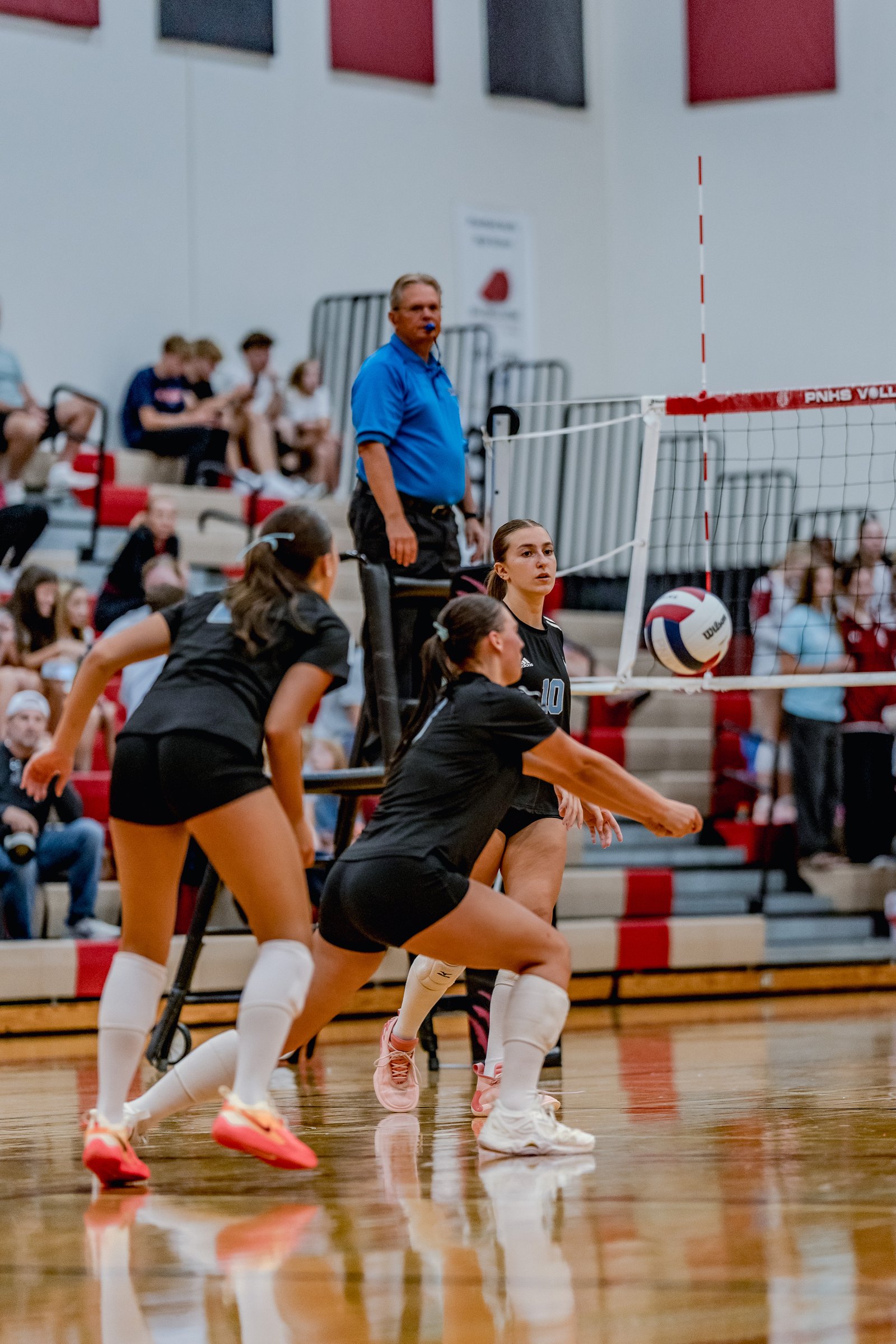 A player in a black top with number 10 passes a volleyball during a game, with teammates and a referee visible.