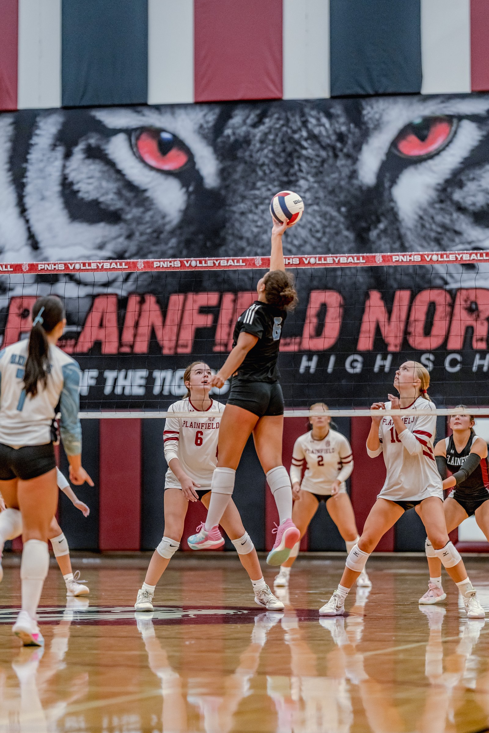 A player in black jumps to hit the volleyball as players in white with maroon trim watch during a game.