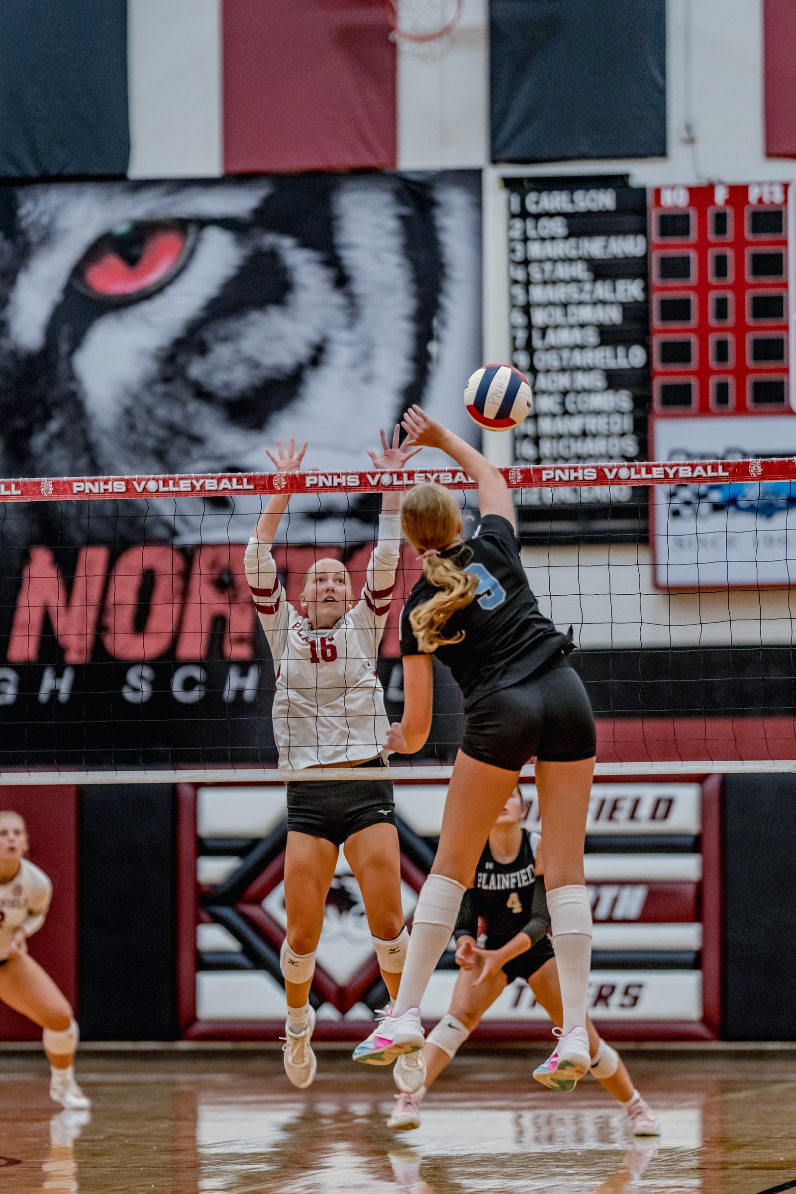 Players in white and black jerseys jump at the net during a volleyball game, with a player in white wearing number 16.