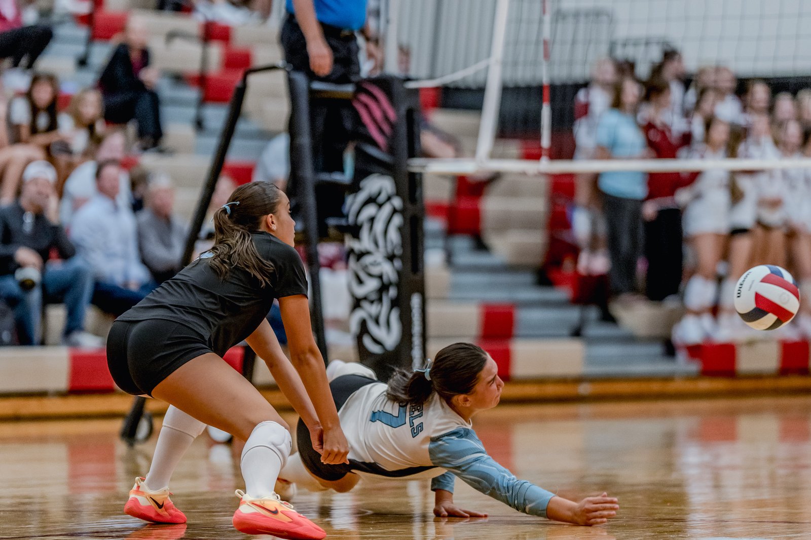 A player in black dives for a volleyball while a player in white and blue attempts a dig on the court with spectators in the background.