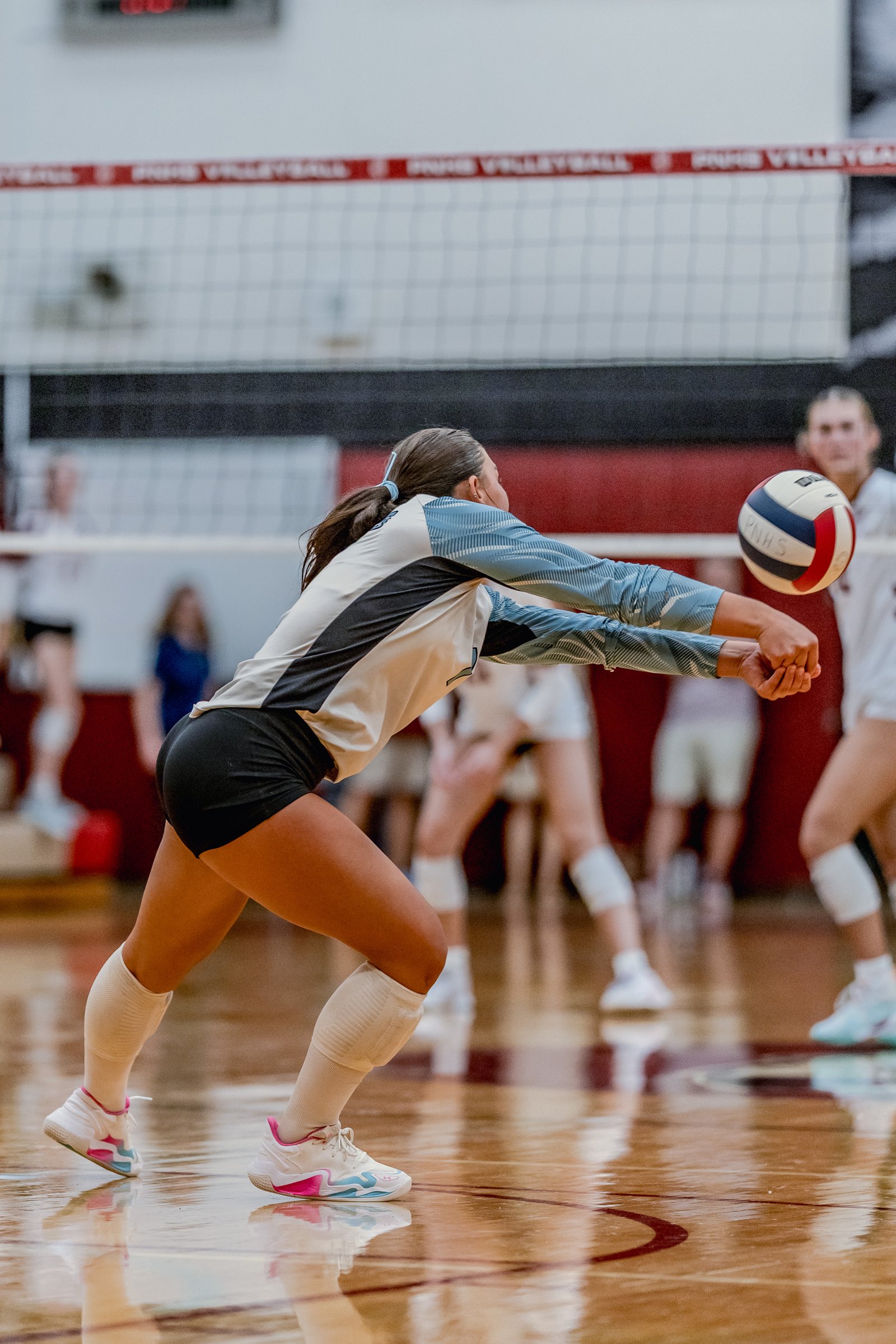 A player in a light blue and black jersey makes a dig on the volleyball court with other players in white jerseys visible.