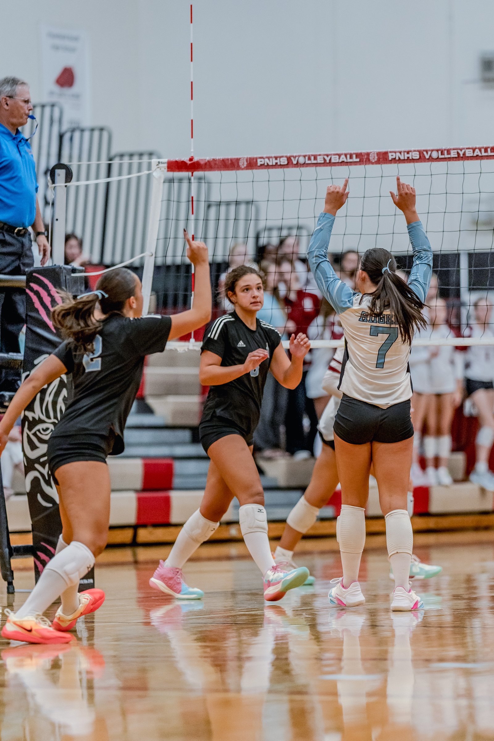 Players in black and white jerseys with blue accents play volleyball, with one player in black raising her hand.