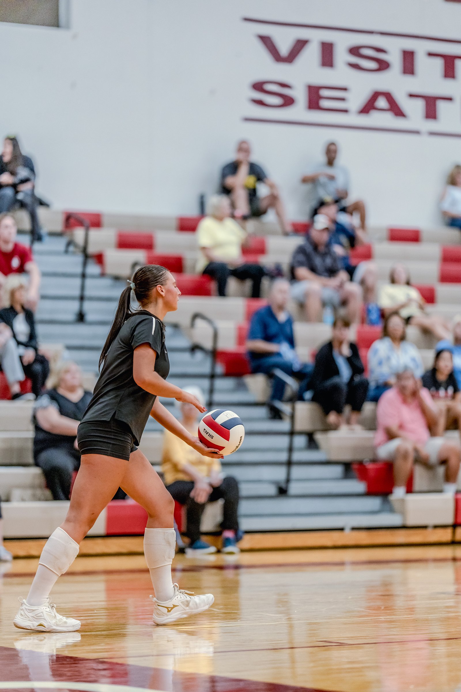A player in a black top and shorts prepares to serve a volleyball during a game, with spectators in the background.