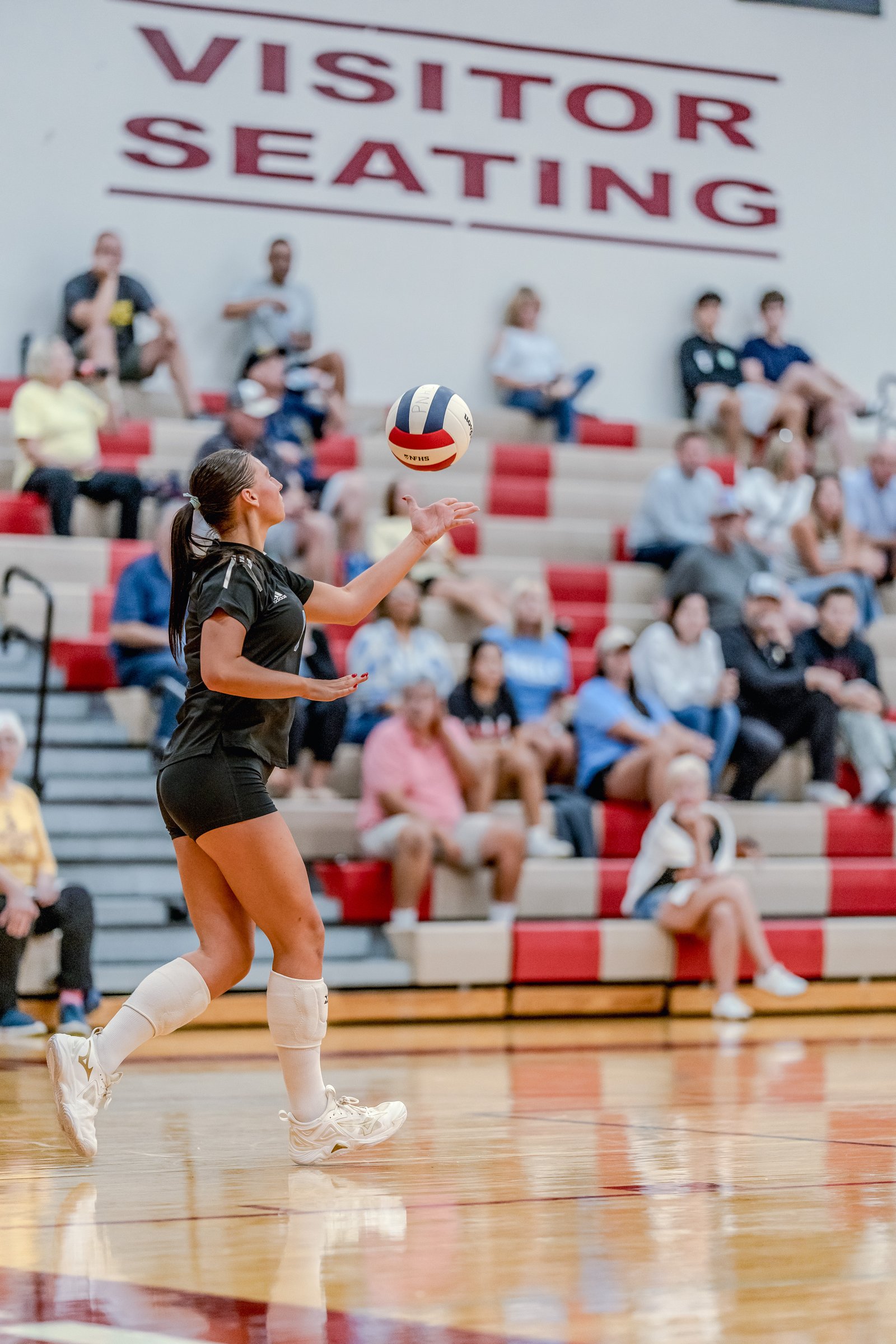 A player in a black top and shorts serves a volleyball in a gymnasium with spectators in the background.