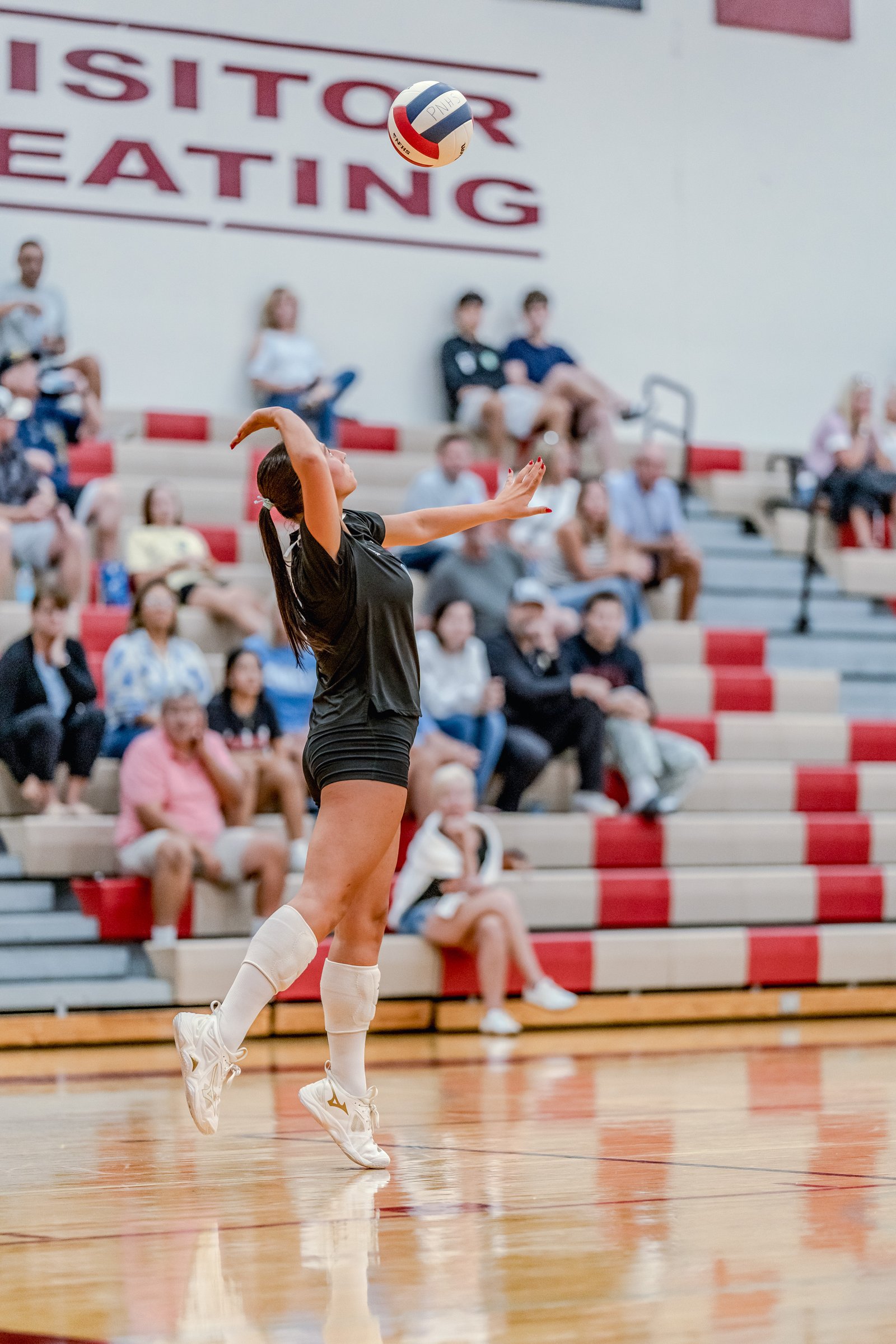 A player in a black top serves a volleyball in a gymnasium with spectators in the background.