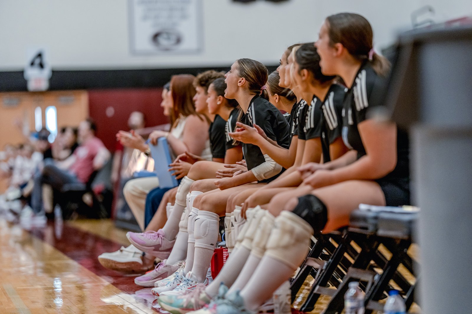 Players in black jerseys with white stripes cheer from the bench during a volleyball game.